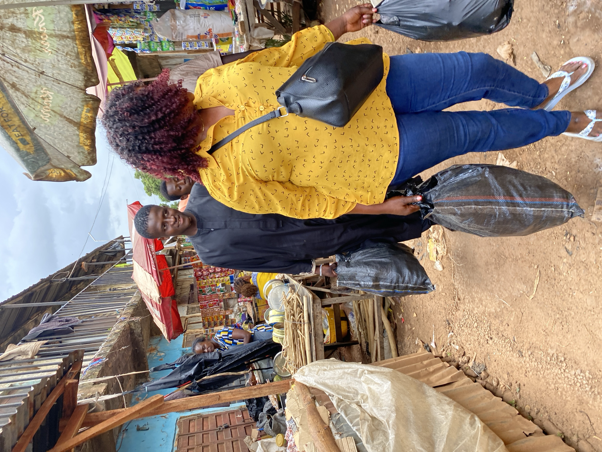 In Bukuru, Nigeria, a street scene unfolds at a market. A Black woman with curly, reddish-brown hair, wearing a yellow, patterned blouse and blue jeans, walks forward, carrying a black handbag over her shoulder and a large black plastic bag in her right hand. To her left, a Black man in a black traditional outfit also walks forward, carrying a similar black plastic bag in his left hand. Behind them, other individuals are visible, including a woman in a blue and yellow patterned dress. The market environment is characterized by stalls with red and white awnings, shelves stocked with goods, and various items on display, including what appear to be food products and household items. Wooden tables hold bundles of sticks and metal containers. The ground is covered in dirt and gravel. The overall atmosphere suggests a bustling, outdoor marketplace.