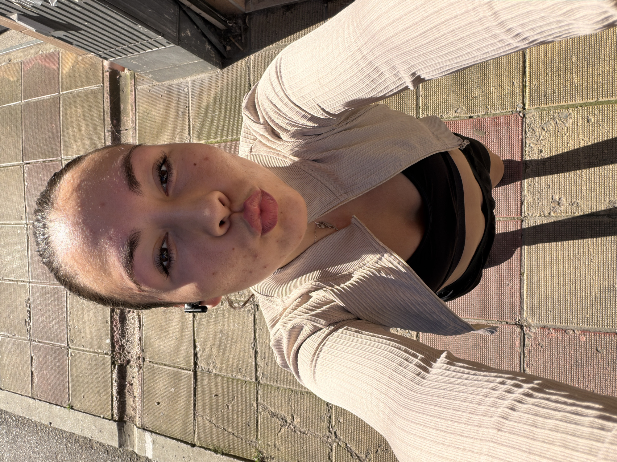 A woman is taking a selfie outdoors in Varna, Bulgaria. She is standing on a sidewalk paved with rectangular concrete tiles in alternating shades of brown and yellow. A shadow of a building falls on the right side of the image, and shadows cast by the woman and her clothing are visible on the tiles. The woman is wearing a light-colored, ribbed cardigan over a black top and dark shorts. She has her hair pulled back and is wearing black wireless earbuds. Her lips are pursed in a kissing gesture, and she is looking directly at the camera. The lighting suggests it is a sunny day.