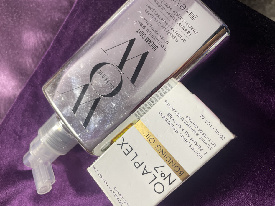 The image shows two hair care products. On the left, a bottle of "COLOR WOW Dream Coat Supernatural Spray", described as a spray prodigieux that transforms texture and provides humidity protection. On the right, a box of "Olaplex No. 7 Bonding Oil", which boosts shine, strengthens, and repairs all hair types. The box indicates a volume of 30ml/1.0 fl. oz. and the bottle is 200 ml. The products are placed on a purple velvet surface.
