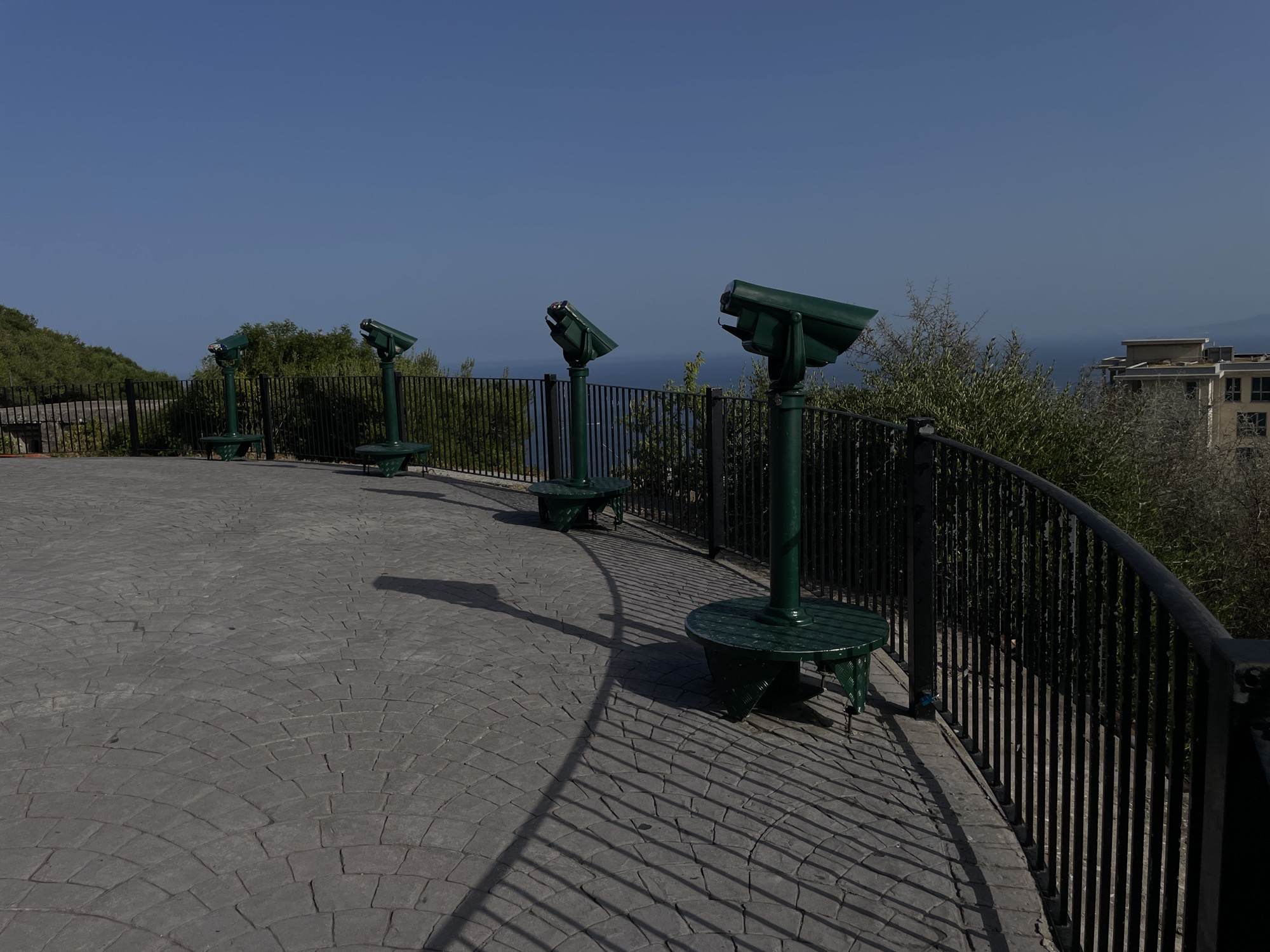 The image shows a scenic viewpoint on the Rock of Gibraltar, a British Overseas Territory. In the foreground, a paved pathway with a distinctive fan pattern leads the eye towards the horizon. Along the pathway, there are four green, metal public binoculars mounted on pedestals, angled towards the sea. A black metal railing lines the edge of the viewpoint, offering safety and a clear view of the expanse beyond. Behind the railing, lush green foliage and trees create a natural barrier, with a glimpse of a modern building in the distance on the right. The sky is a clear, brilliant blue, and the overall impression is one of a well-maintained tourist attraction offering breathtaking panoramic views.