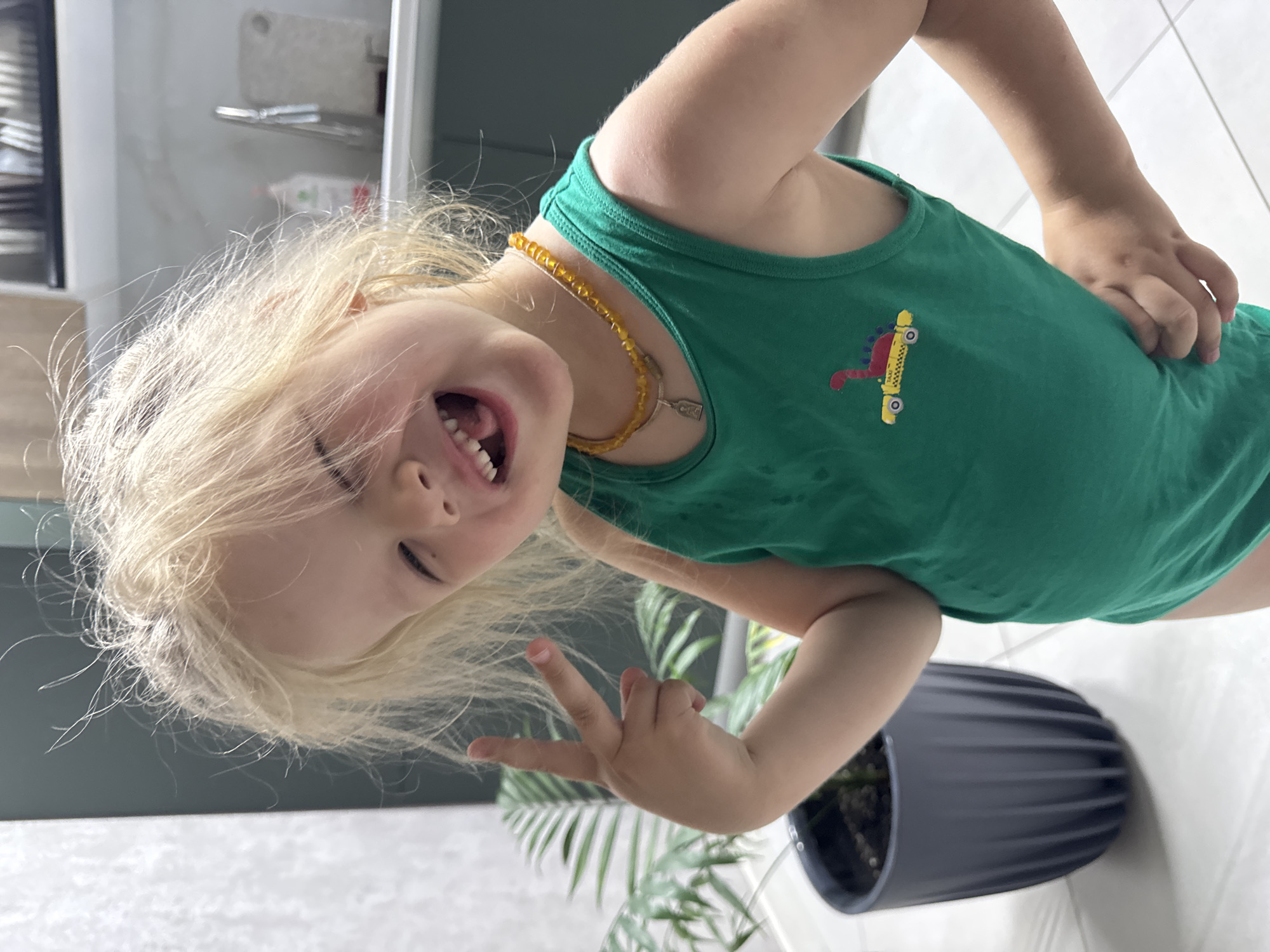 A young child with blond hair is captured in a distorted angle. The child wears a green tank top with a yellow and red taxi design. They are wearing a beaded amber necklace. They are positioned near a plant in a black pot, and the background suggests an indoor environment with a refrigerator or cabinet. The location mentioned, Ферма, Russia, cannot be definitively confirmed as the location based on this image alone. The child is making a peace sign with their fingers and showing their teeth.