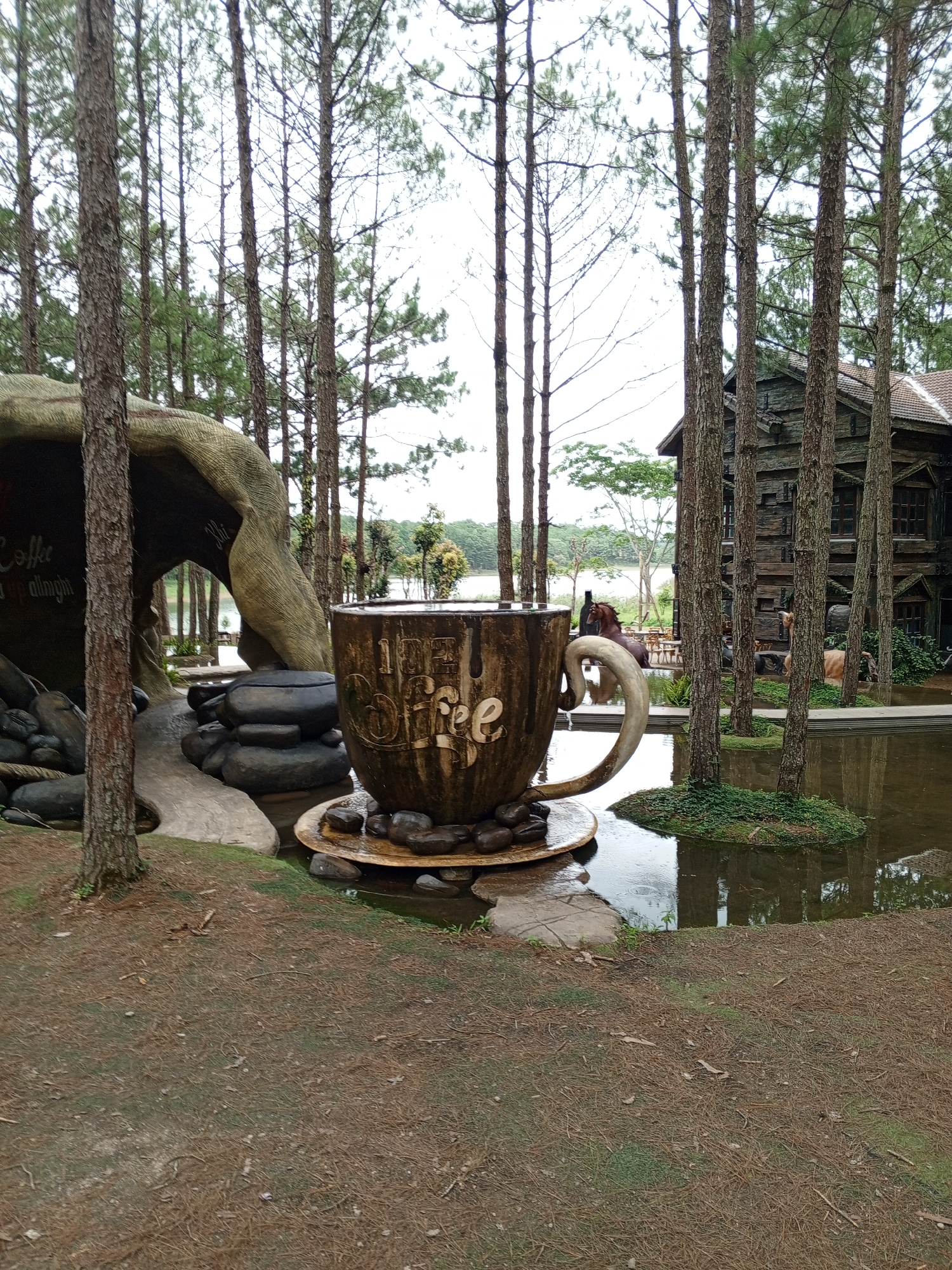 The image captures a scene at "Thung Lũng Tình Yêu" (Valley of Love) in Da Lat, Vietnam. In the foreground, a large, stylized coffee cup sculpture sits on a saucer made of wood and stone. The cup is weathered and appears to be made of wood, with the word "Coffee" prominently inscribed on its side. The sculpture is surrounded by water and a small patch of greenery. To the left, a large, abstract rock formation curves into a cave-like structure, with what appears to be more stones and a path leading into it. In the background, a rustic wooden house with dark siding and multiple windows stands amongst tall pine trees. Beyond the house, a body of water, likely a lake, is visible, with trees and distant hills forming the horizon. Two horses, one brown and one lighter in color, are present near the water's edge. The overall environment suggests a park or resort setting, with a focus on natural elements and whimsical artistic installations. The lighting indicates an overcast day.
