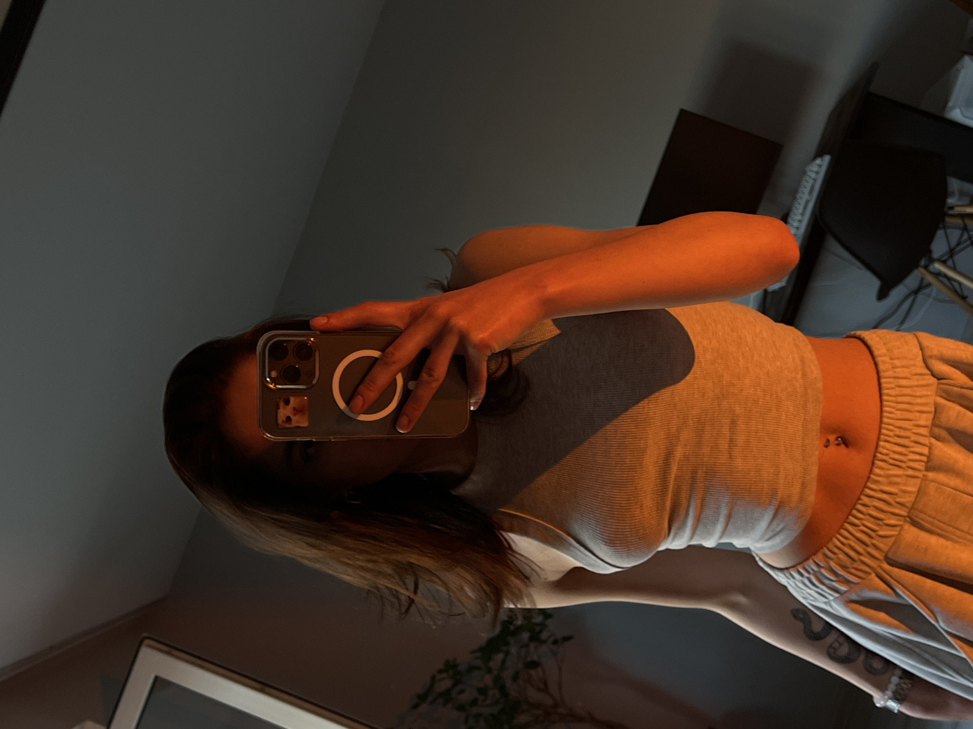 A young woman is taking a mirror selfie in a dimly lit room. She is wearing a grey ribbed tank top and light grey sweatpants with a drawstring waist. A silver belly button ring is visible. She has a tattoo on her left arm, which appears to be a snake. She is holding a smartphone in her right hand, capturing her reflection. The lighting in the room is warm and orange-toned, possibly from a nearby light source, casting shadows. In the background, a mirror reflects the scene, as well as a glimpse of a plant and a framed object. A desk with a computer monitor and keyboard is also visible. The location is identified as Ternopil, Ukraine.