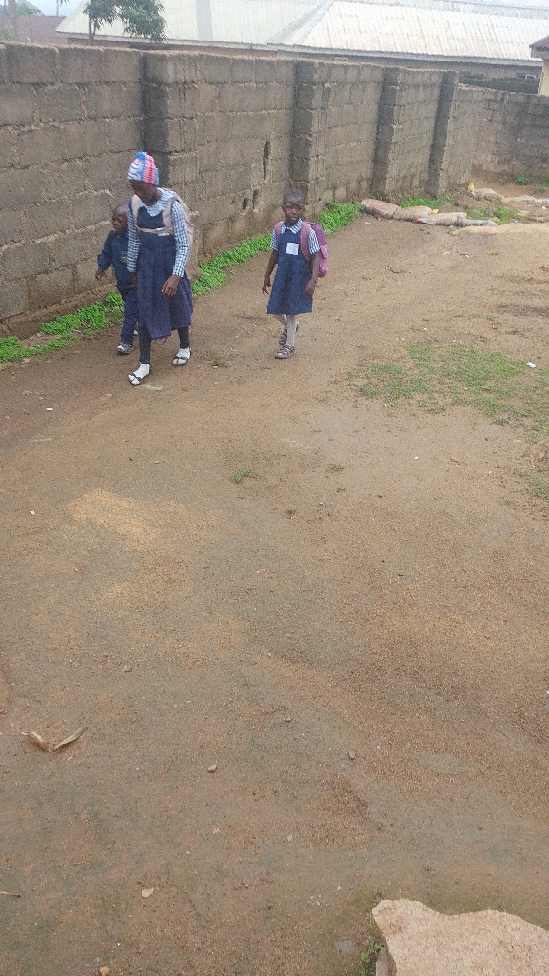 Three children, two girls and one boy, are walking on a dirt path in front of a brick wall. The girl on the left is wearing a blue dress and a striped headscarf, and is carrying a pink backpack. The girl on the right is also wearing a blue dress and a pink backpack. The boy is wearing a blue shirt and pants. The environment appears to be a residential area with a building in the background. The image was taken in Jos, Nigeria.