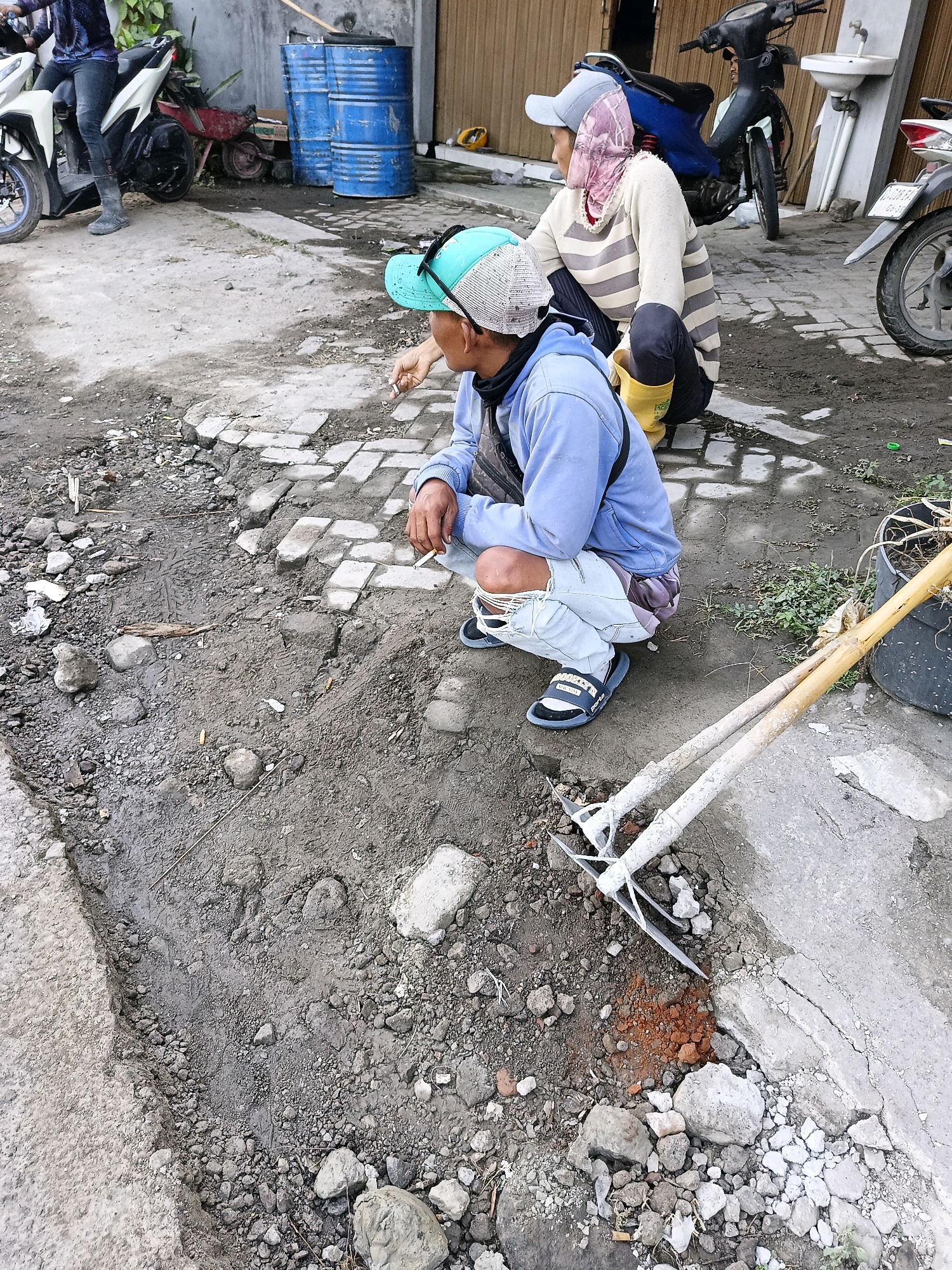 The image depicts an outdoor scene in Sukorini, Indonesia, likely a residential or street area undergoing some form of work or maintenance. In the foreground, a person wearing a light blue hooded sweatshirt, ripped light-colored jeans, and dark slide sandals is crouched down. They are holding a lit cigarette in their right hand. To the right of this individual, a gardening tool with two pointed metal ends and wooden handles lies on the ground, resting against a concrete surface. The ground itself is a mix of dirt, gravel, and broken brick or concrete, suggesting a construction or repair site.
Further back, another person is also crouched, wearing a light-colored striped long-sleeved shirt, dark pants, and yellow boots. They are wearing a light blue baseball cap and a patterned headscarf. Behind them, a dark-colored motorcycle is parked. In the background, two large blue barrels are visible, and part of a white motorcycle is seen to the far right. The environment appears somewhat unkempt, with scattered debris and vegetation. The overall impression is one of everyday life and possibly manual labor in a localized Indonesian setting.