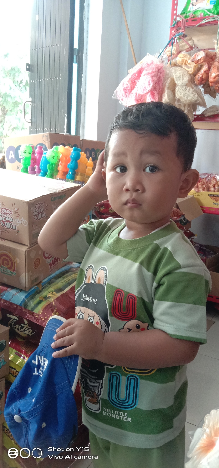 The image depicts a young child, approximately 2-4 years old, standing indoors in what appears to be a small shop or storage area in Pesawahan, Indonesia. The child is positioned centrally in the frame, facing directly towards the camera with a neutral expression.
The child has dark, short, wavy hair. He is wearing a short-sleeved t-shirt with alternating horizontal light green and white stripes. The t-shirt features a graphic print on the front: a cartoon character resembling a rabbit with large ears, the text "Labubu!" above it, a large red stylized letter "U" to the right, and the text "THE LITTLE MONSTER" near the bottom right of the graphic, next to a small brown creature. Light green shorts or pants are visible below the t-shirt.
The child's left hand is raised, with fingers lightly touching the back of his head/ear. His right hand holds a blue fabric baseball cap, which hangs downwards, showing partially visible white embroidered text "ST VER" and a white button on the cap's crown.
The background is cluttered with various items. To the child's left, a stack of cardboard boxes is visible, with one box displaying "OKKY Scan & Win" and a bicycle logo, and another marked with "A B C" letters. On top of these boxes, a row of colorful plastic figurines or bottles (green, pink, blue, orange, yellow) are lined up.
To the child's right and above his head, several clear plastic bags containing various packaged snacks, likely crackers or chips, are hung from a red metal shelving unit. The snacks are predominantly pink, white/beige, and orange. In the far left background, a dark-colored, multi-paneled accordion-style door or security shutter is partially visible, adjacent to a bright white wall or column. A slender, light-colored pole leans against the wall in the upper right background. The overall lighting suggests an interior space, possibly illuminated by natural light from an opening.