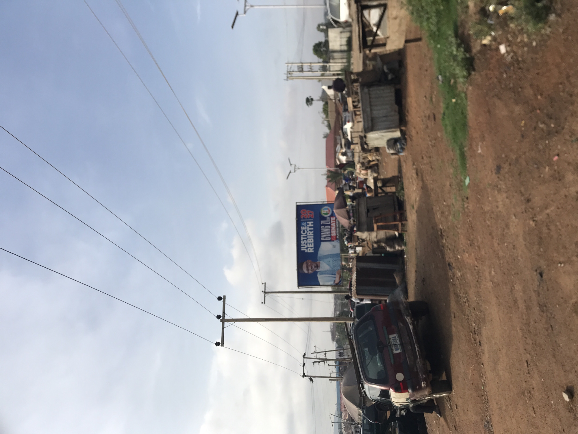 This daytime image captures a roadside scene in Rafin Par, Nigeria, characterized by a mix of infrastructure, commerce, and political messaging. The frame is horizontally split, with a pale blue, partly cloudy sky dominating the upper left and a dusty, reddish-brown dirt road filling the lower right.
Along the left side of the road, multiple wooden utility poles rise, supporting a dense network of black power lines that crisscross the sky. Some poles also hold streetlights. In the foreground, on the right side of the dirt road, a red minivan or SUV is parked, facing away from the viewer.
Further down the road, on the left, a large blue billboard is prominently displayed. It features the portrait of a man and carries the text "JUSTICE & REBIRTH 2023 GYANG ZL SENATE PLATFORMNP". Beyond the billboard and stretching into the midground, various informal structures, stalls, and buildings line the roadside, hinting at commercial activity. A few indistinct figures are visible near these structures. The ground beside the road shows some scattered debris and patches of green vegetation. The overall atmosphere suggests a busy, yet somewhat informal, area with bright daylight conditions.