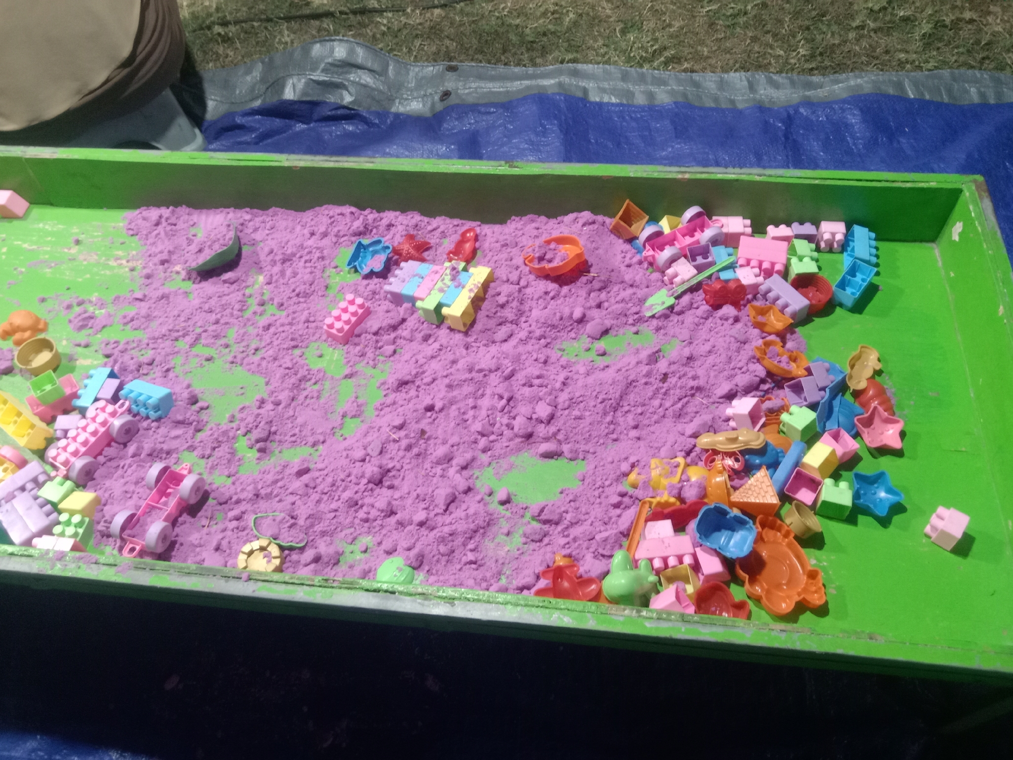 The image depicts a green sandbox filled with purple kinetic sand. Numerous colorful plastic toys are scattered throughout the sand, including building blocks, toy cars, molds in various shapes (stars, fish, shells), and other small construction pieces. The sandbox itself is situated outdoors, appearing to be on a blue tarp which is placed on a grassy area. The background shows more grass and a dark, out-of-focus area. There are no people visible in the image. This scene is a typical setup for children's play.