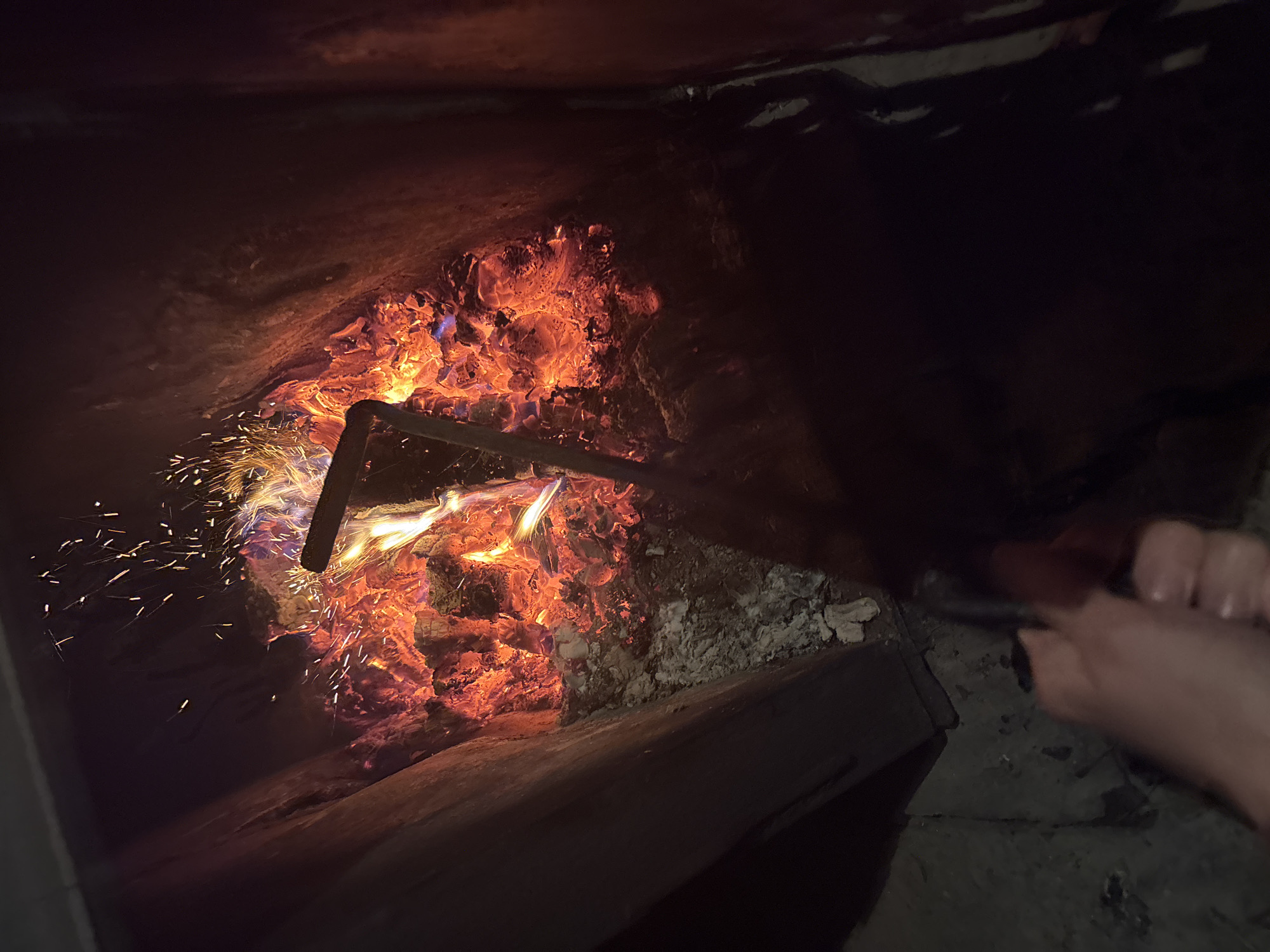 A close-up, indoor shot shows a fire burning in what appears to be a fireplace or furnace. The flames are bright orange and yellow, with sparks rising upwards. A dark, metallic poker tool is partially submerged in the coals. To the lower left, a blurred hand is visible, holding a dark object, possibly a lighter or a part of the poker. The surrounding structure is dark and appears to be made of stone or brick, with some ash and debris on the floor. The scene suggests a cold environment where a fire is being maintained for warmth. The location in the city of Курган, Russia is not specified by any visual cues in the image.