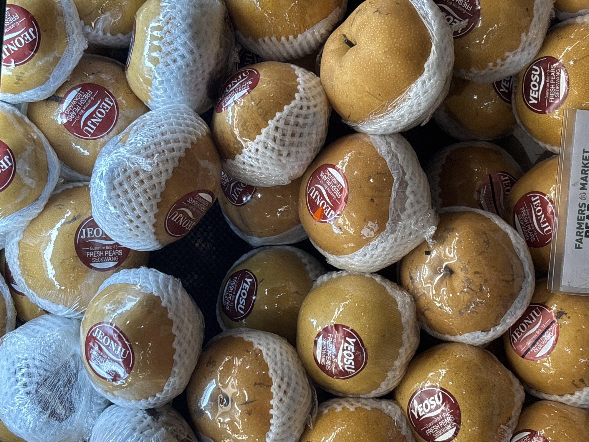 The image displays a close-up, overhead view of a large pile of Asian pears, likely in a market stall. The pears are golden-brown in color, individually wrapped in clear plastic, and cushioned by white protective netting. Many of the pears have red circular stickers with white text. Some stickers read "JEONJU" and "FRESH PEARS SEOKWANG," while others say "YEOSU" and "FRESH PEARS SEOKWANG," suggesting they are from different regions or brands. The pears are stacked in a somewhat disorganized manner, with some partially obscured. The lighting is somewhat dim, consistent with an indoor market setting. The image does not contain any explicit visual cues to determine the specific location within North Jakarta, Indonesia, beyond the general context of a fruit market. There are no people or discernible actions visible in the frame.