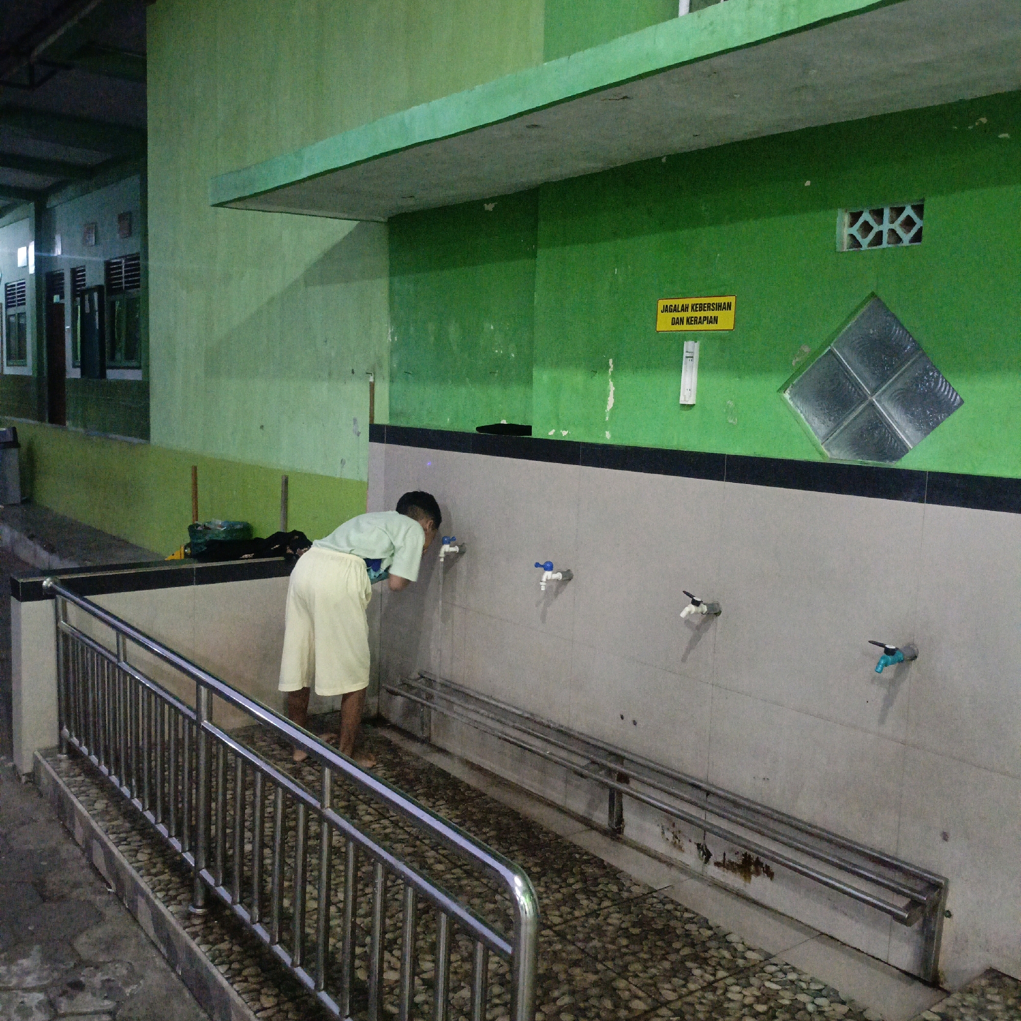 This image depicts an outdoor area with a green wall and a tiled wall, likely part of a public facility or institution in Salatiga, Indonesia. In the foreground, a metal railing separates a walkway from the tiled area. A young person, wearing a light green t-shirt and pale yellow shorts, is bending down to wash their face at a faucet on the tiled wall. Water is running from the faucet. Several other faucets are visible along the wall, some with blue handles and others with black and white handles. On the green wall, a yellow sign with black text reads "JAGALAH KEBERSIHAN DAN KERAPIAN," which translates to "Maintain Cleanliness and Tidiness." There is also a decorative vent or window with a diamond pattern on the green wall. The ground is paved with small stones and some darker square tiles. In the background, there is a building with windows and a covered walkway. Some bags or items are placed on the ground near the tiled wall.