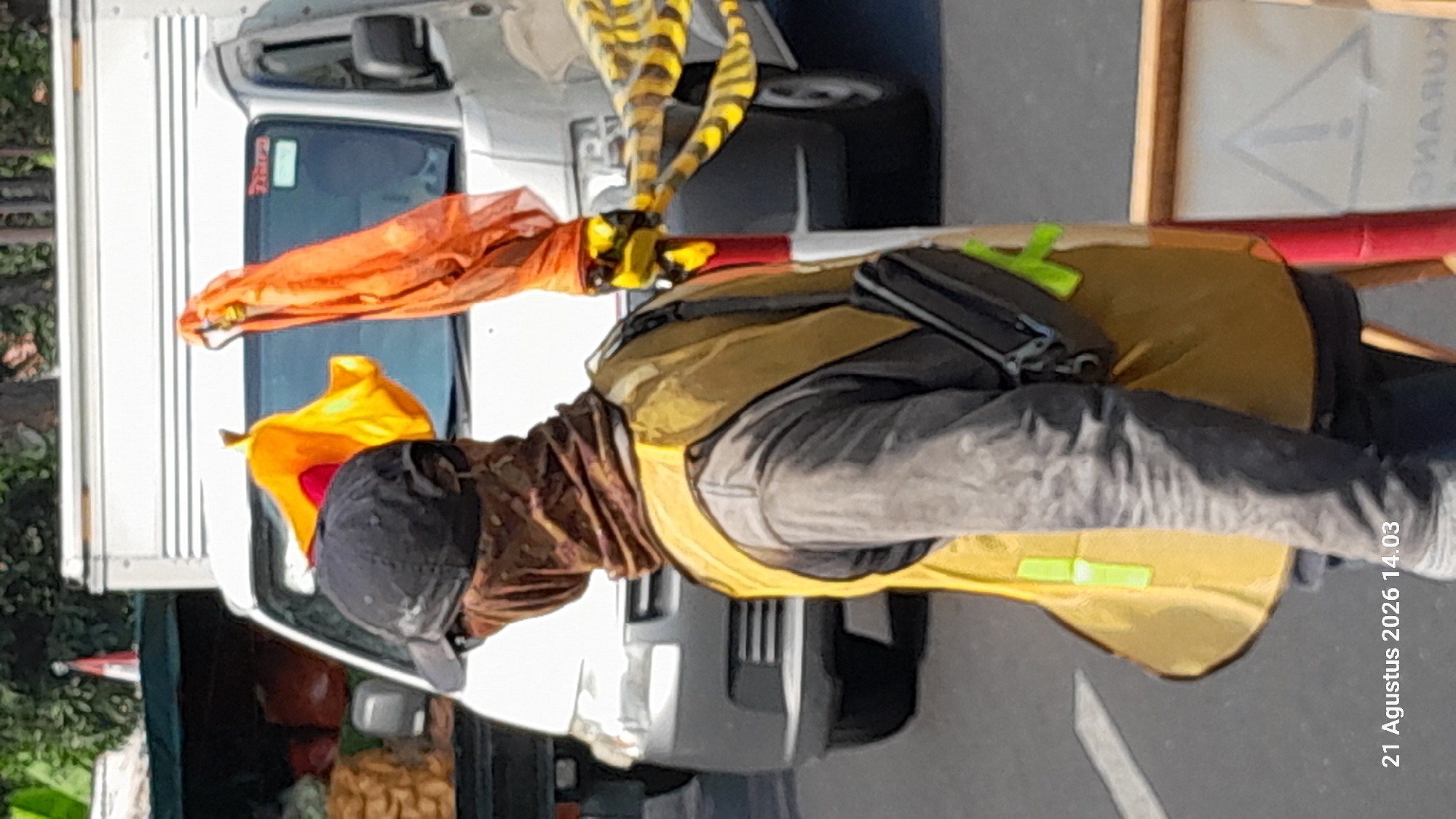 A person wearing a dark baseball cap, a patterned face mask covering their nose and mouth, and a yellow safety vest over a grey long-sleeved shirt stands on a street. They are facing away from the camera, looking towards a white truck. The truck has a red sign on the windshield and appears to be parked. In front of the truck, there is a pole with orange and yellow fabric draped over it. Yellow and black striped caution tape is also visible, wrapped around the pole and extending to the right. To the left of the truck, part of a dark green vehicle or stall with produce bags is visible. The street has a white line marking. In the bottom right corner, a sign with an exclamation mark inside a triangle is partially visible. The timestamp "21 Agustus 2026 14.03" is present in the bottom left corner, indicating the date and time. The background shows greenery and trees. This scene is taking place in Takeran, Indonesia.