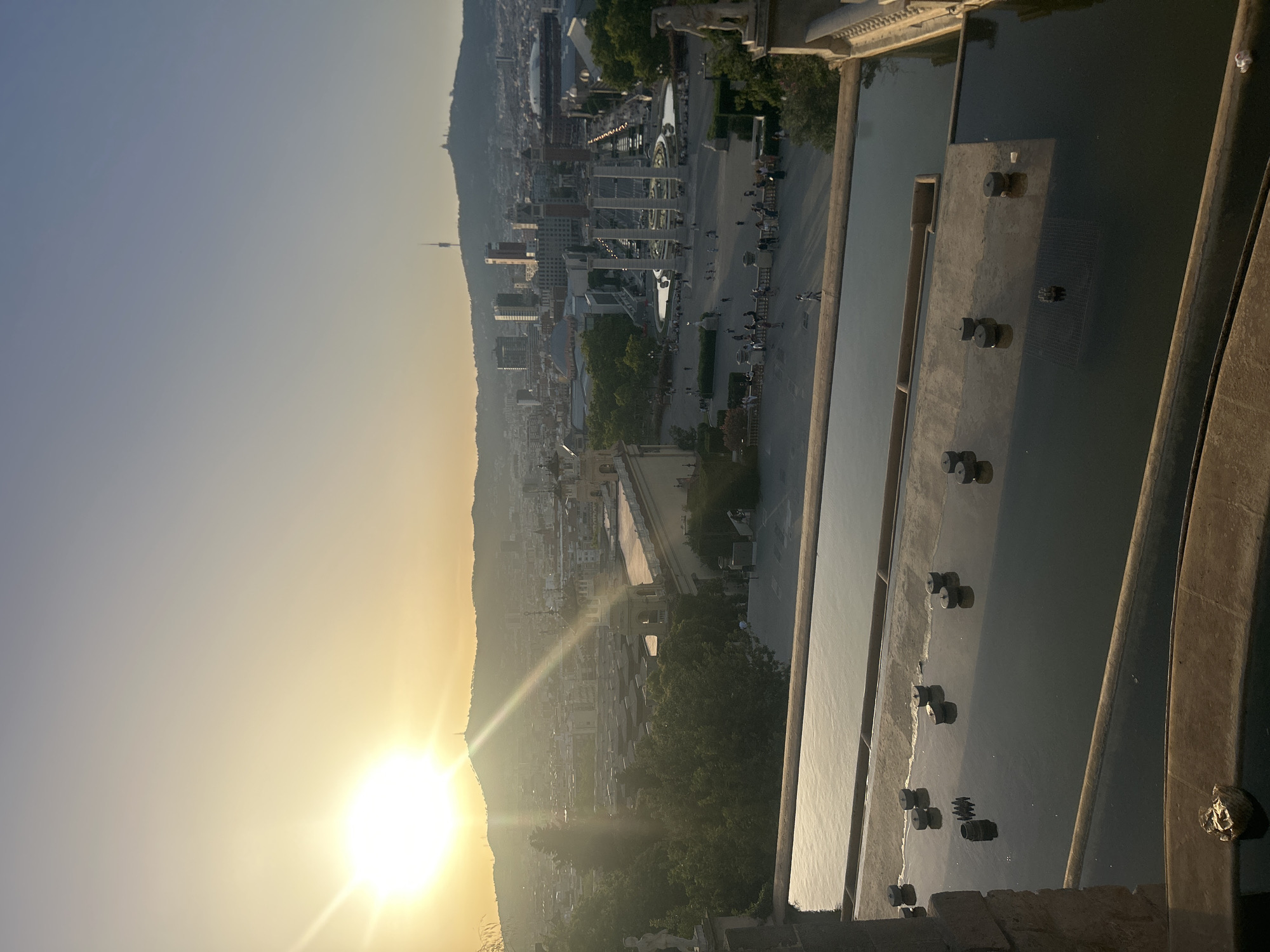The image captures a panoramic view of Barcelona, Spain, during a sunset. The sun is setting behind a mountain range, casting a warm glow over the city. In the foreground, there are tiered water features, likely part of a grand fountain system, with several circular elements submerged in the water. Beyond the water features, a cityscape unfolds, featuring modern buildings and older architectural structures. A prominent landmark in the middle ground is a large plaza with a central fountain surrounded by columns and several people. The overall impression is of a serene urban landscape bathed in the soft light of dusk.