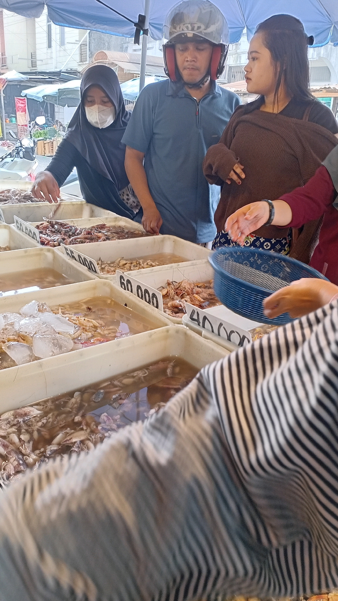 The image depicts a bustling fish market scene in Jambi, Indonesia, likely located in a street market or a dedicated fish trading area. The foreground is dominated by a person wearing a black and white striped shirt, partially obscuring several large, shallow trays filled with various seafood. These trays are submerged in water, and some contain ice. Visible seafood includes shrimp and what appear to be small squid or cuttlefish. Price tags are visible on some of the trays, with prices like "60.000" and "40.000" indicated.
In the mid-ground, three individuals are interacting near the seafood stalls. A woman wearing a black hijab and a white face mask is reaching into one of the trays, appearing to select seafood. Beside her, a man wearing a blue polo shirt and a motorcycle helmet is looking towards the woman. To the right of the man, a woman wearing a brown knitted top is looking slightly away from the camera, with her left hand holding a blue plastic basket and her right hand gesturing. Another person, wearing a dark red sleeve, is partially visible on the far right, also interacting with the basket.
The background reveals the open-air market environment. A blue tarp is stretched overhead, providing shade. Stalls with various goods and awnings are visible further back, along with what appears to be a motorcycle parked to the left, suggesting a typical urban market setting in Indonesia. The overall impression is one of active commerce and daily life in a local Indonesian market.
