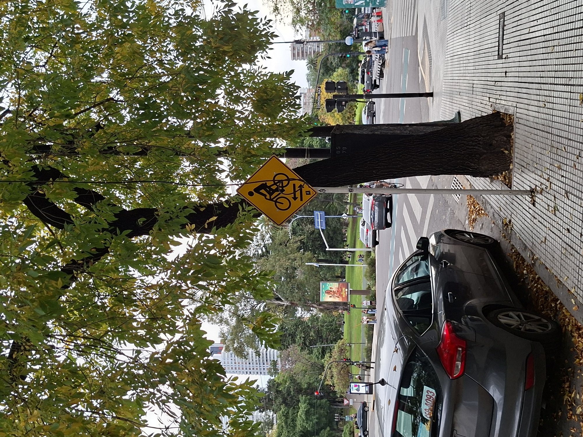 The image depicts a street scene in Buenos Aires, Argentina, evidenced by the "ARS" license plate visible on a gray hatchback car parked along the curb. The sidewalk is tiled with a gray and white grid pattern. A tree trunk obscures part of a yellow diamond-shaped traffic sign indicating a bicycle crossing with a pedestrian. Beyond the tree and sign, a park with green grass and trees is visible, with distant buildings. People can be seen in the background walking and standing on the sidewalk and in the park. Traffic lights and cars are seen on the street. Based on the street signs, it's likely the intersection of Coronel Diaz and Gral. Justo José de Urquiza.