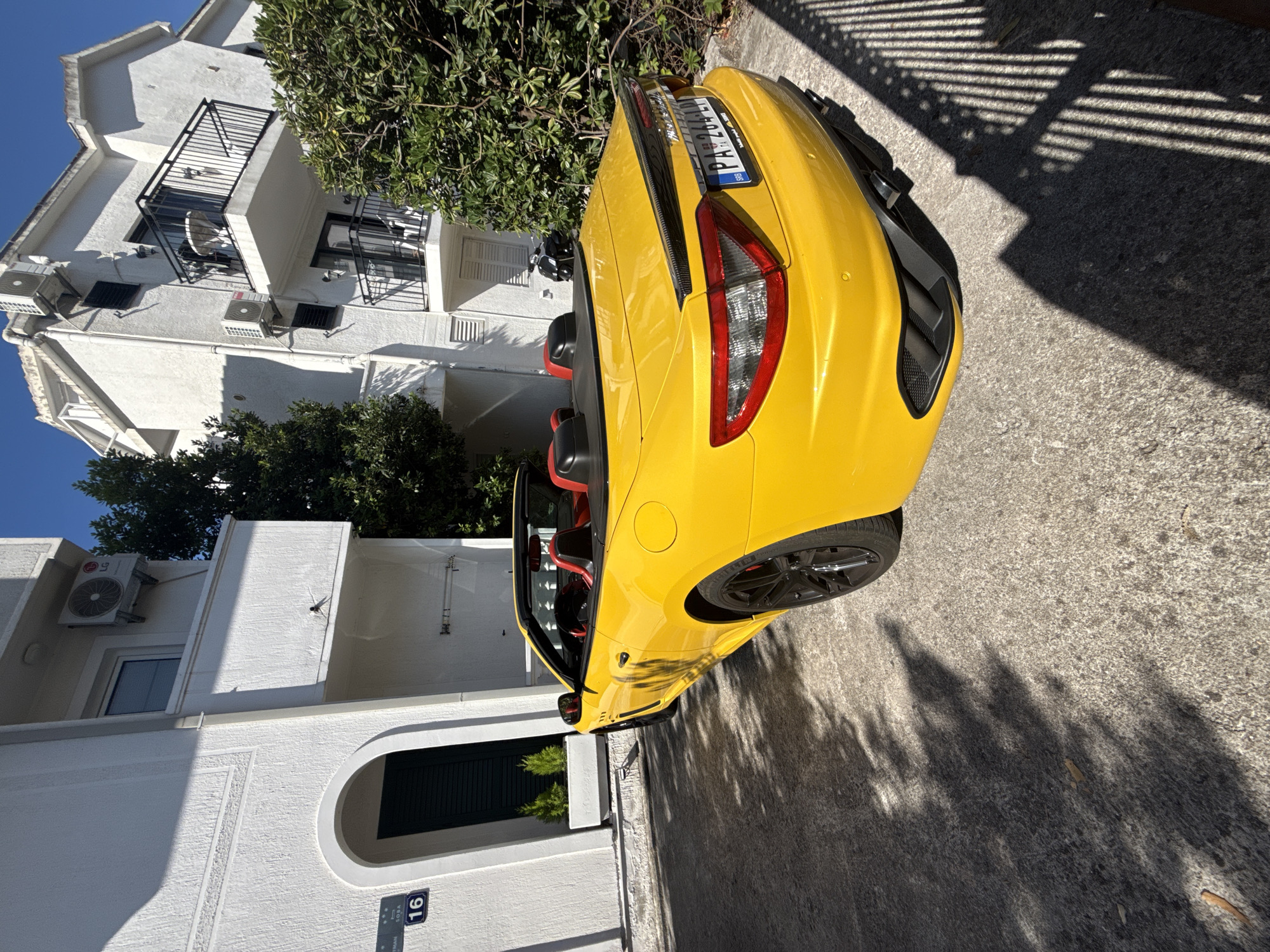 A bright yellow Maserati GranTurismo convertible is parked on a concrete driveway in front of a white stucco building. The car's interior is visible, featuring black seats with red accents. A license plate with the characters "PA 264 LP" is mounted on the rear of the vehicle. The building has multiple windows and balconies, and an air conditioning unit is mounted on one of the walls. The scene is bathed in sunlight, casting strong shadows on the ground. A black motorcycle is parked to the right of the car, partially obscured by foliage. The environment appears to be residential, possibly in Budva, Montenegro, given the presence of what appears to be a street sign with the number "16" and the text "SOBA," which translates to "room" in Montenegrin, suggesting a guesthouse or apartment rental.