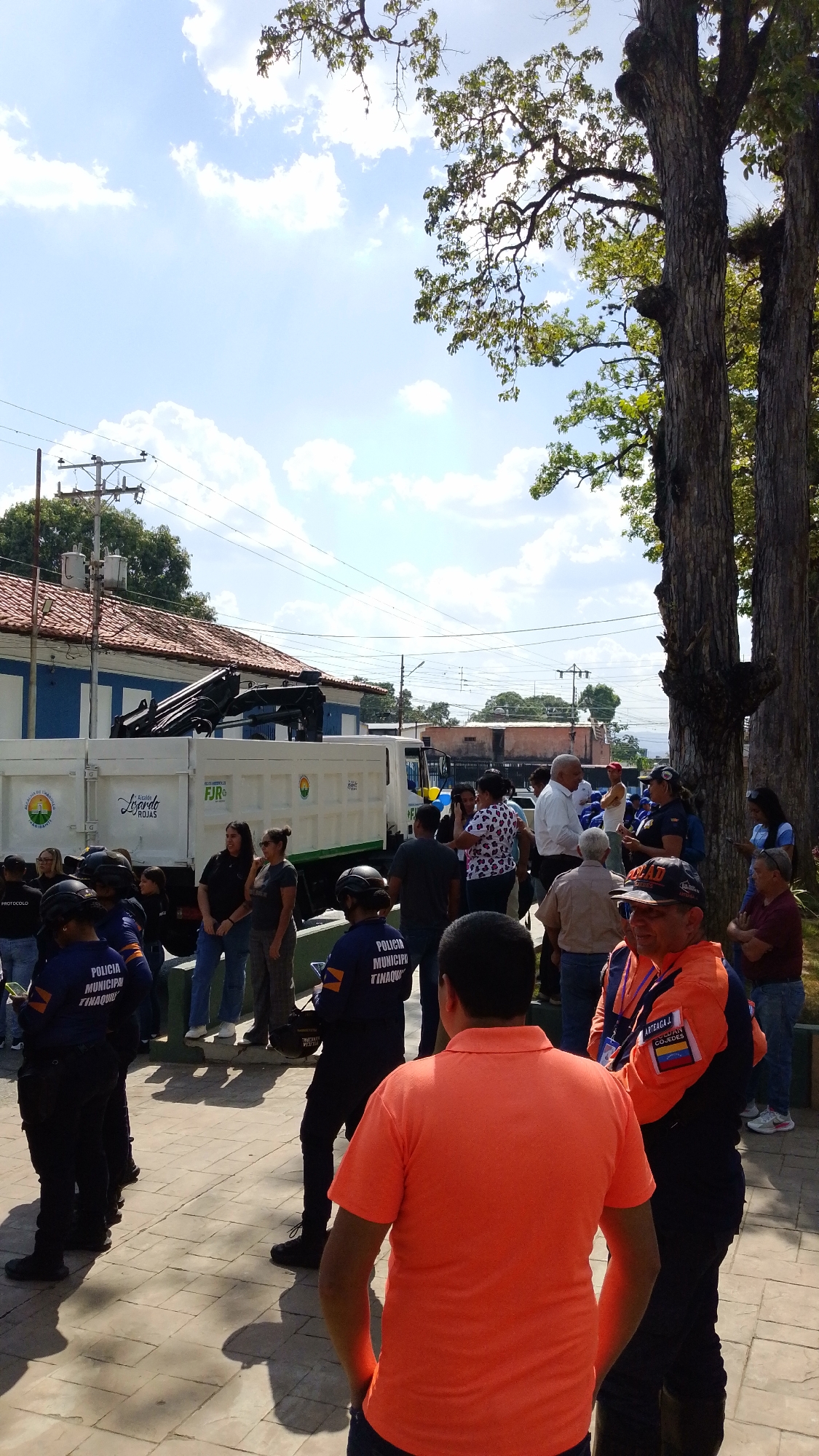 A sunny day in Tinaquillo, Venezuela. A crowd has gathered on a paved plaza beside a white utility truck, labeled “FJR” and displaying the Tinaquillo municipality logo. The crowd comprises civilians and uniformed personnel, some of whom are identified as “POLICIA MUNICIPAL TINAQUILLO”. Other uniformed people are in orange jackets that say "ARTEAGA" and "COJEDES" with a Venezuelan flag. A two-story building with a red tiled roof sits in the background, and mature trees provide partial shade. Power lines are visible overhead.