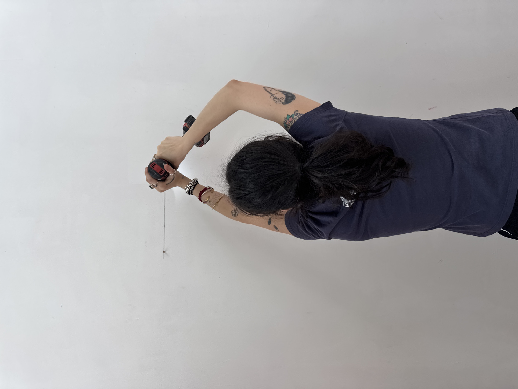 A person wearing a dark-colored t-shirt is using two red and black power drills on a white wall. A thin chain hangs from the wall near the lower drill. The location appears to be indoors, potentially in the Town of Red Hook, United States of America.