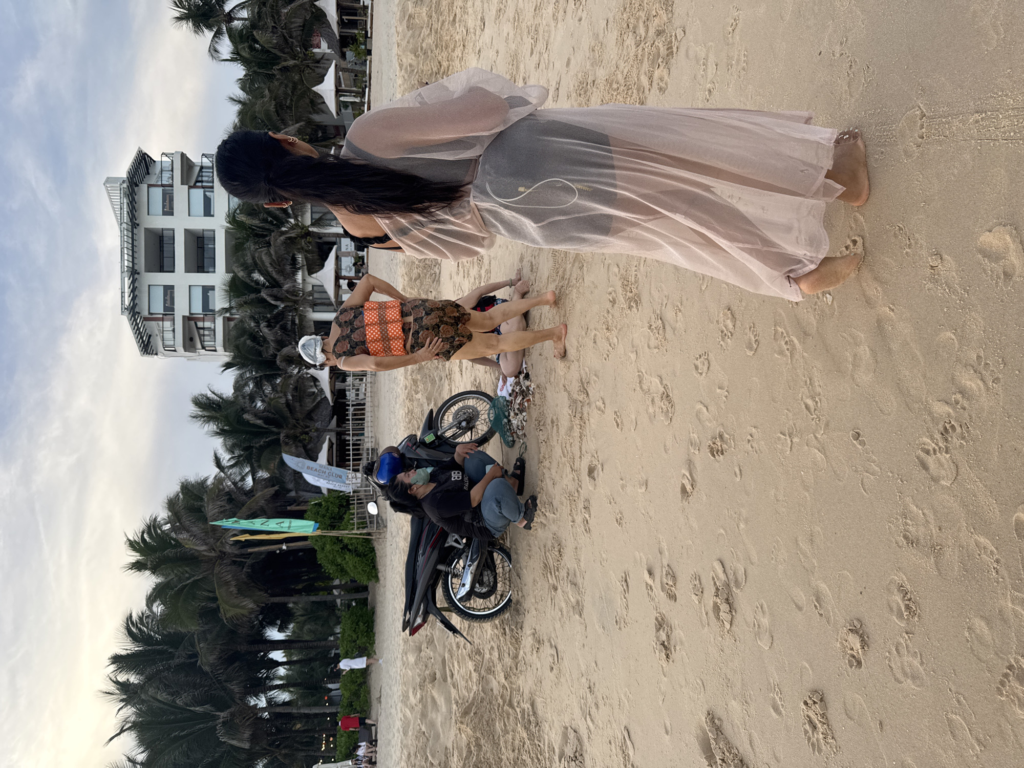 The image depicts a beach scene in Phu Thuy Ward, Vietnam. In the foreground, a person wearing a sheer, light pink robe stands facing away from the camera, their back to the viewer. Their dark hair is pulled back in a ponytail. To the left and further back, another person is crouching next to a parked motorcycle. This person is wearing a black t-shirt and blue jeans, with a light green mask partially covering their face. The motorcycle is dark in color with red accents. In the mid-ground, two more individuals are visible. One is wearing a patterned top and shorts, and another person's legs are visible lying on the sand. A group of people are seated or standing near some tents and palm trees in the background, with a multi-story white building visible beyond the trees. The environment is a sandy beach with scattered palm trees, flags, and beach umbrellas. The sky is overcast.
