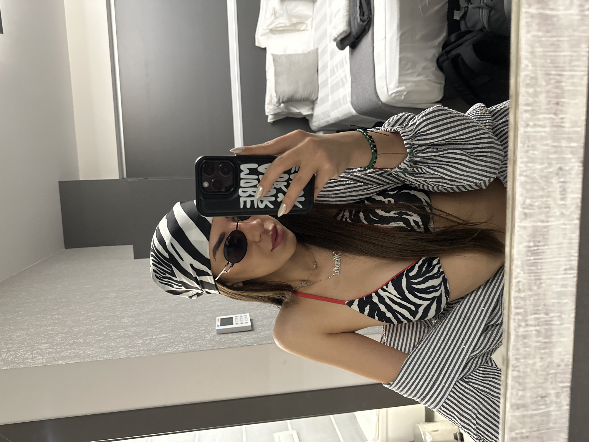 A young woman with long dark hair, wearing a zebra-print bikini top, a black and white striped shirt unbuttoned, a zebra-print headscarf, and oval sunglasses, takes a mirror selfie in what appears to be a hotel room in the city of Ofrynio Beach, Greece. She is holding up a smartphone with a black case that has white lettering. She is also wearing a necklace and bracelets. In the background, a bed with white linens and a gray throw is visible, along with luggage on the floor. The room has gray walls and textured wallpaper.