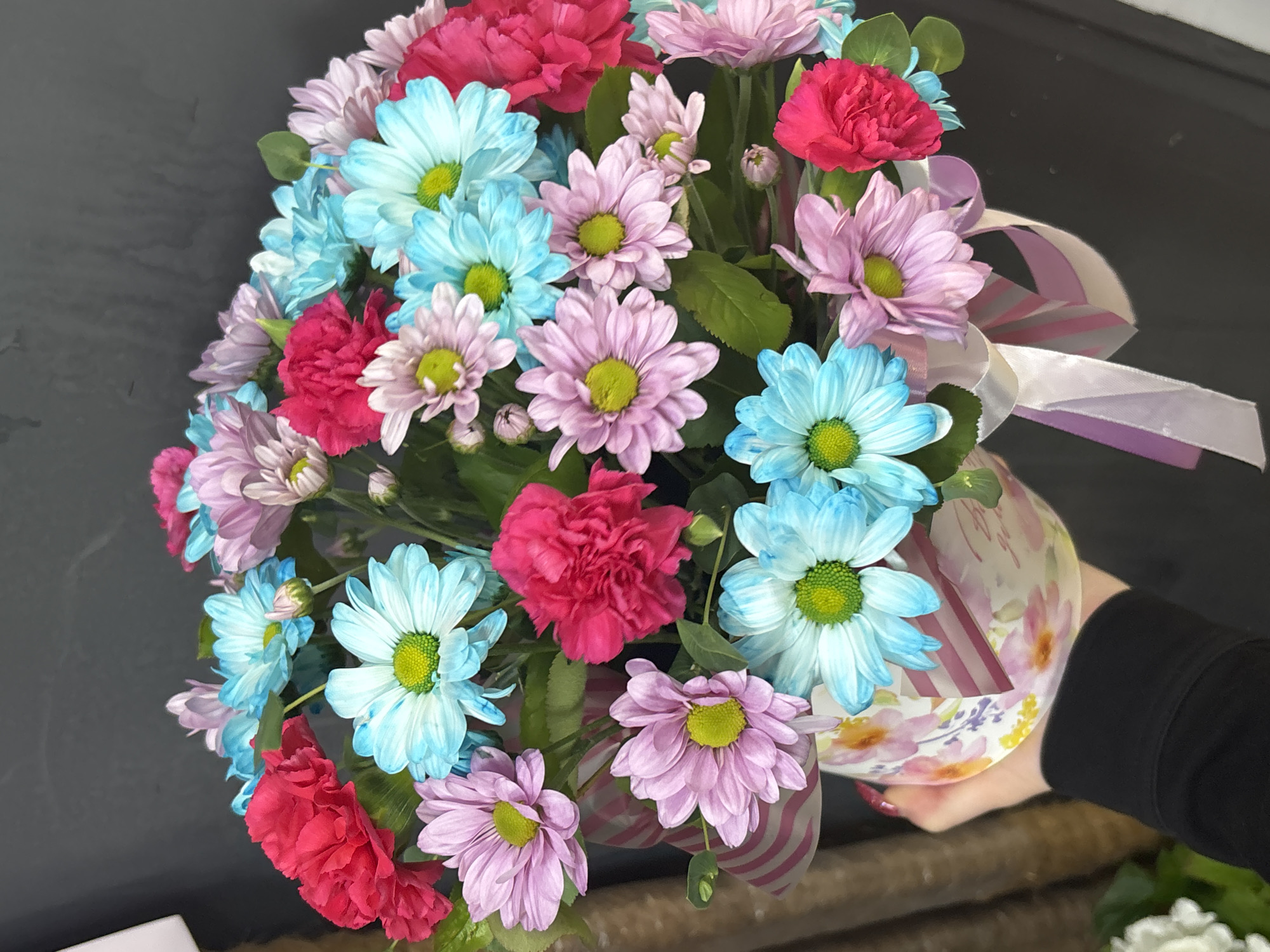 A bouquet of bright, colorful flowers is held by a person's hand. The bouquet features a variety of daisy-like flowers in shades of pink, light blue, and magenta carnations. The flowers are arranged in a decorative patterned box adorned with pink and white striped ribbon and a white bow. The background is dark and out of focus, with a hint of what appears to be a dark wall. The person's hand is covered by a black long-sleeved shirt.