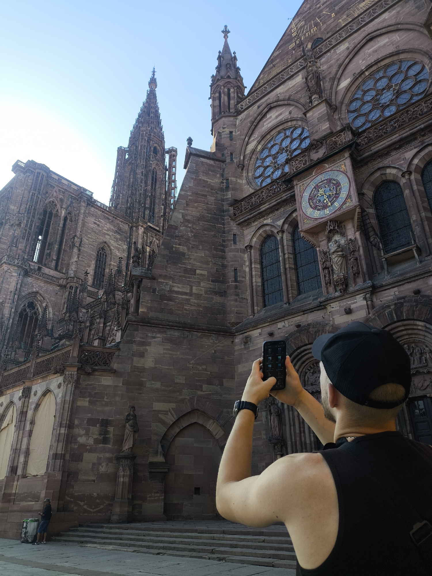 The image shows a person holding a smartphone to take a picture of the Strasbourg Cathedral, also known as the Cathédrale Notre-Dame de Strasbourg, in Strasbourg, France. The cathedral is a Gothic masterpiece, characterized by its towering spires and intricate stonework. The front of the cathedral features large rose windows and a detailed astronomical clock. The person in the foreground is wearing a black tank top and a black baseball cap, facing away from the camera. The sky is clear and blue. The scene captures a moment of tourism and appreciation for historical architecture.