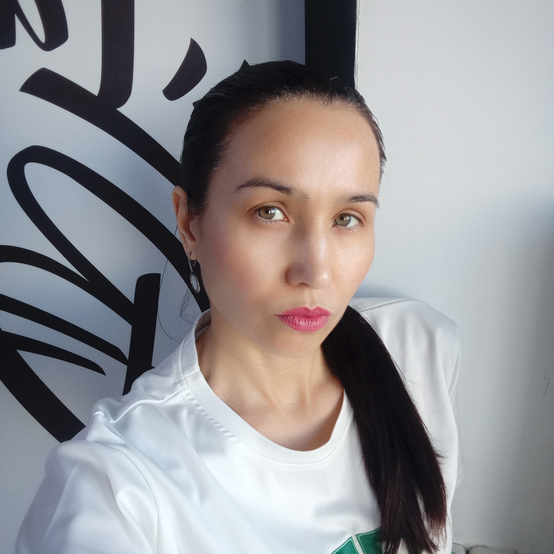 A medium close-up selfie shows a woman with dark hair pulled back in a ponytail, wearing a white t-shirt with a green design visible on the front. She has fair skin, light brown eyes, and pink lipstick. An earring hangs from her left ear. Behind her are a white wall and a section of a graphic with black, thick, curved lines resembling calligraphy. The location is unspecified, and no information about Izhevsk, Russia, is present.