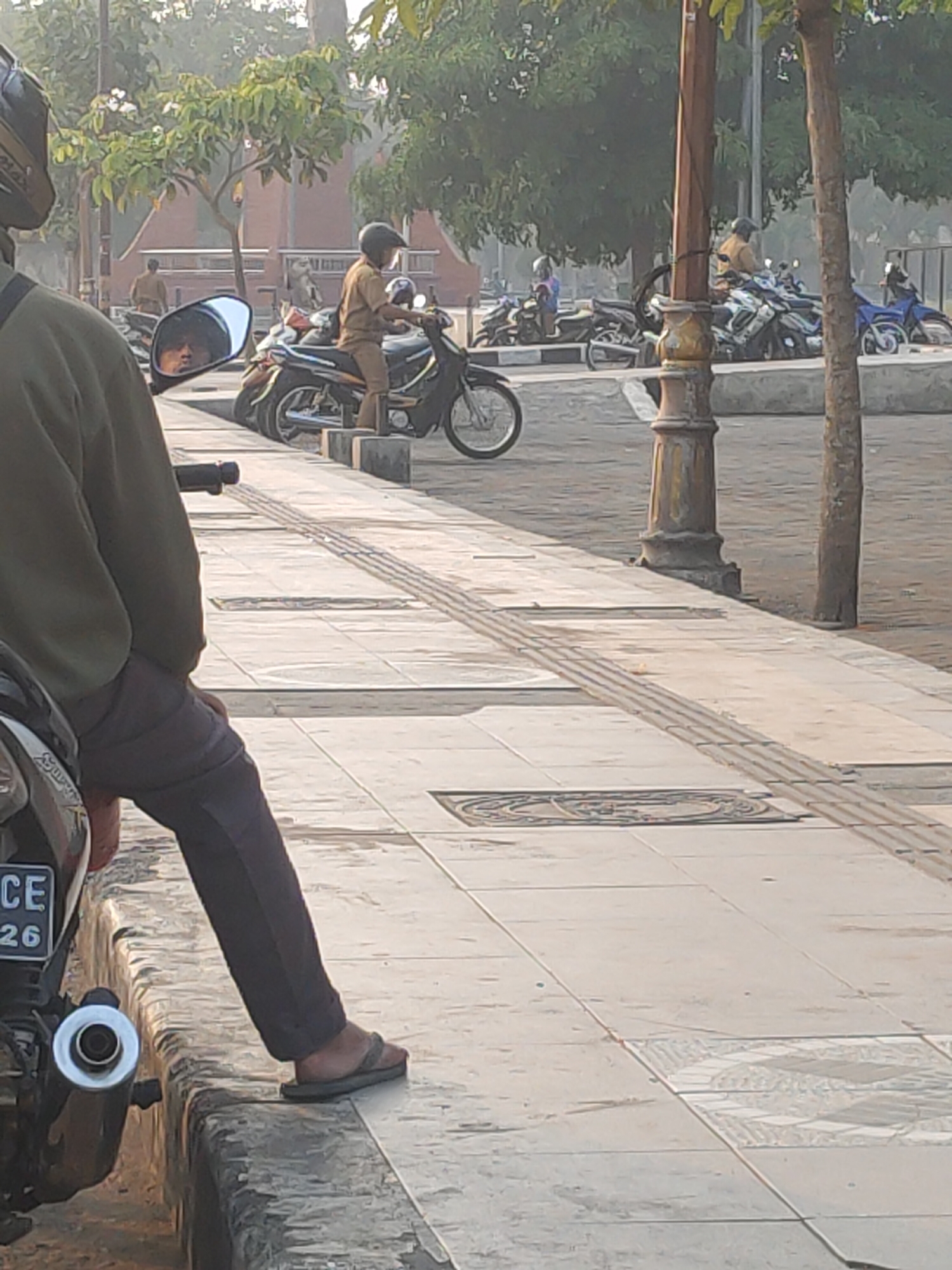 The image captures a street scene in Kanigoro, Indonesia, during daylight hours. In the foreground, a person is seated on a motorcycle, facing away from the camera. The motorcycle has a license plate with the characters "CE 26". To the left, another motorcycle is partially visible. In the middle ground, a person in a tan uniform and helmet is seated on a black motorcycle. Several other motorcycles are parked behind this individual. The background features trees and a hazy sky. The ground is paved with tiles, and a textured strip runs along the edge of the walkway. A lamppost is also visible.