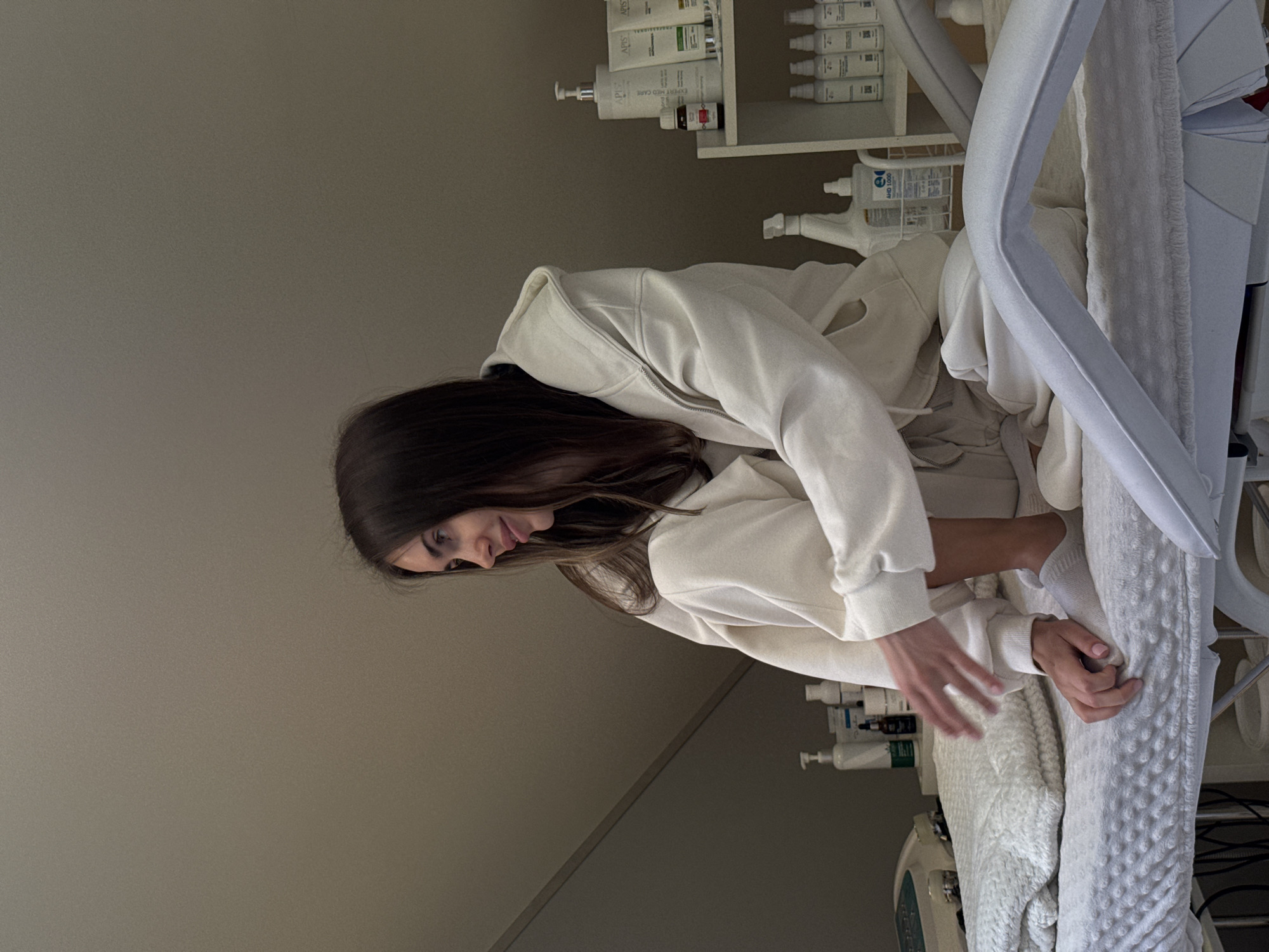 A woman in a white hoodie is next to a medical bed. Shelves stocked with lotions and other medical products are behind her. A machine is visible near the base of the bed. The location is likely a medical or cosmetic clinic.