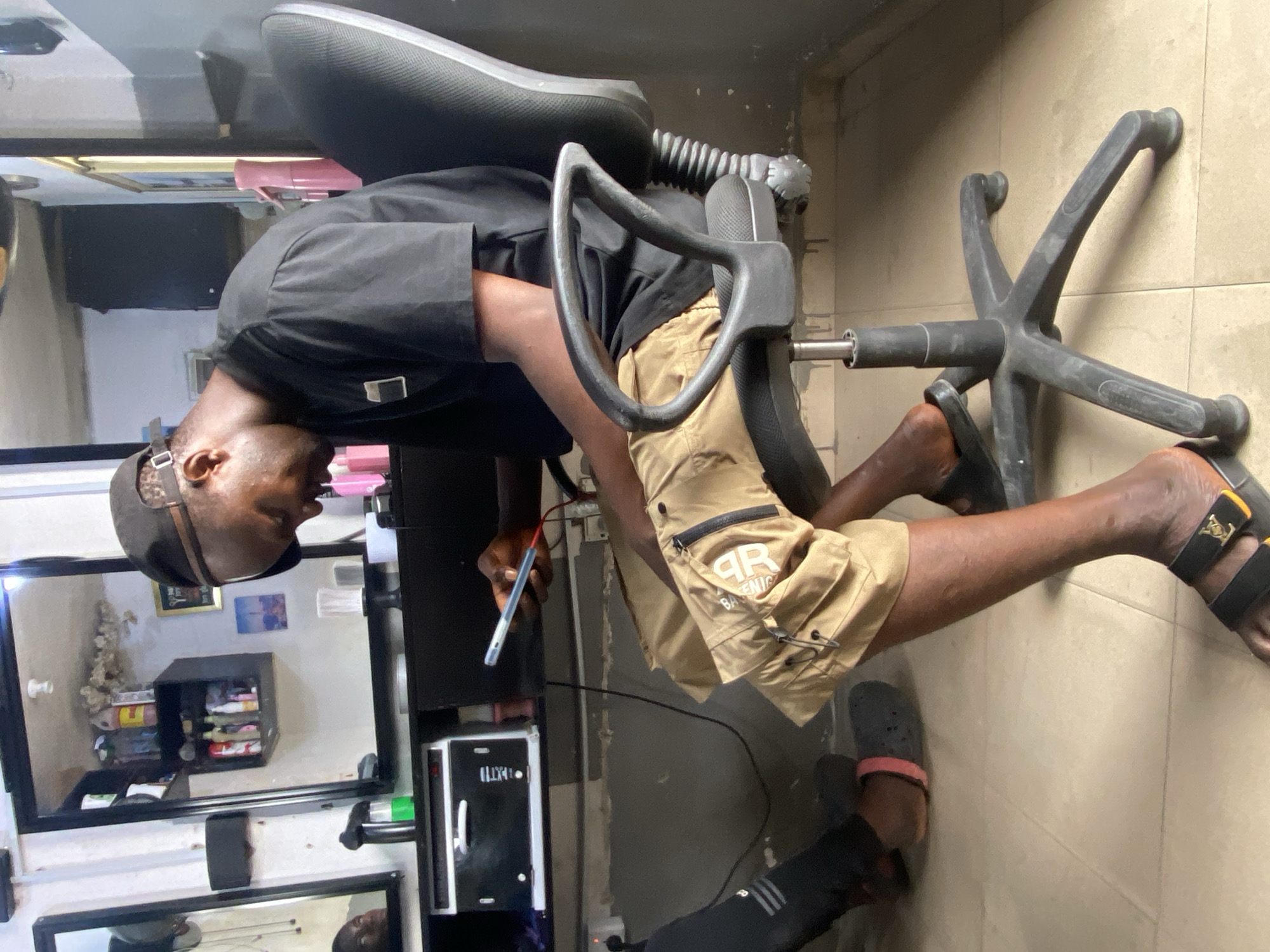 A male individual is captured in an indoor setting, seated on an office chair with a detached wheel base. He is looking down at a smartphone held in his right hand, which appears to be connected to a charging cable.
The man has dark skin and is wearing a black cap, a black short-sleeved shirt, and tan cargo shorts. On his right foot, he wears a black sandal with a gold buckle, while on his left foot, he has a black and pink slide. A logo "AR BASENIG" is visible on the pocket of his shorts. Gold letters, possibly "LV", are discernable on the sandal strap.
The chair he is sitting on has a black seat and backrest, with the main hydraulic cylinder visible as the caster base is disconnected and lies on the tiled floor beside him. The background reveals a room with light-colored tiled floors and walls. Multiple mirrors are affixed to the walls, reflecting parts of the room, including a black square shelf unit holding various items, and a smaller black cabinet below it. Wires are visible on the floor around the chair. The overall scene suggests a casual, possibly waiting, moment within what could be a salon or a personal workspace in Ado, Nigeria.