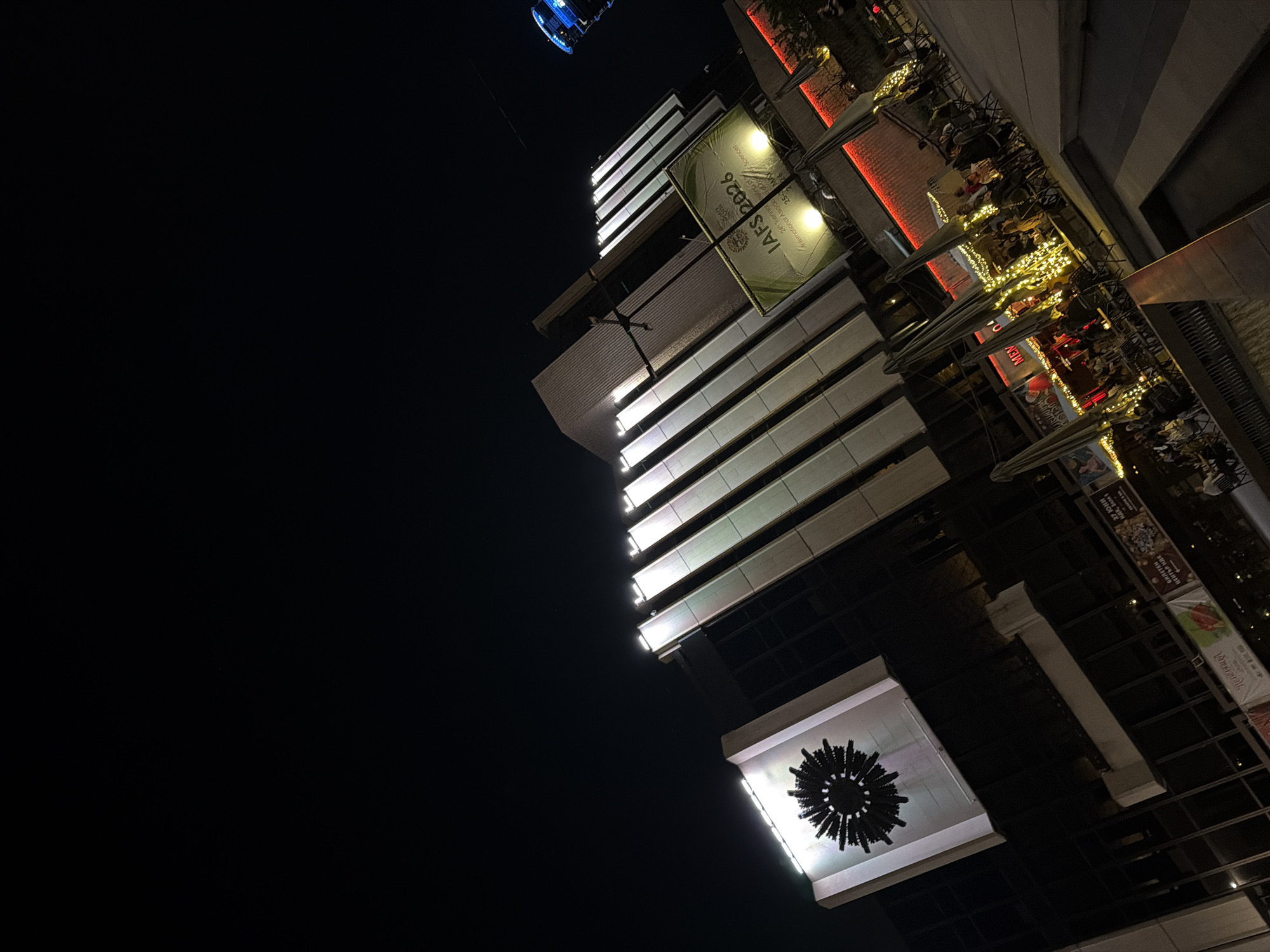 Captured at night, the image shows an urban scene with a building prominently featured. The building has a sign that reads "IAFS 2026", and parts of the text "International Association" and "Sciences" are visible. There's an outdoor area that looks like a restaurant or café with tables and umbrellas, and the area is illuminated with string lights. Additional signs and displays can be seen on the building, one partially reading "БИЛЕТЕН ЦЕНТЪР НДК". A round, decorative object with a starburst-like pattern is affixed to the building, and a faint circular structure appears in the upper left corner.