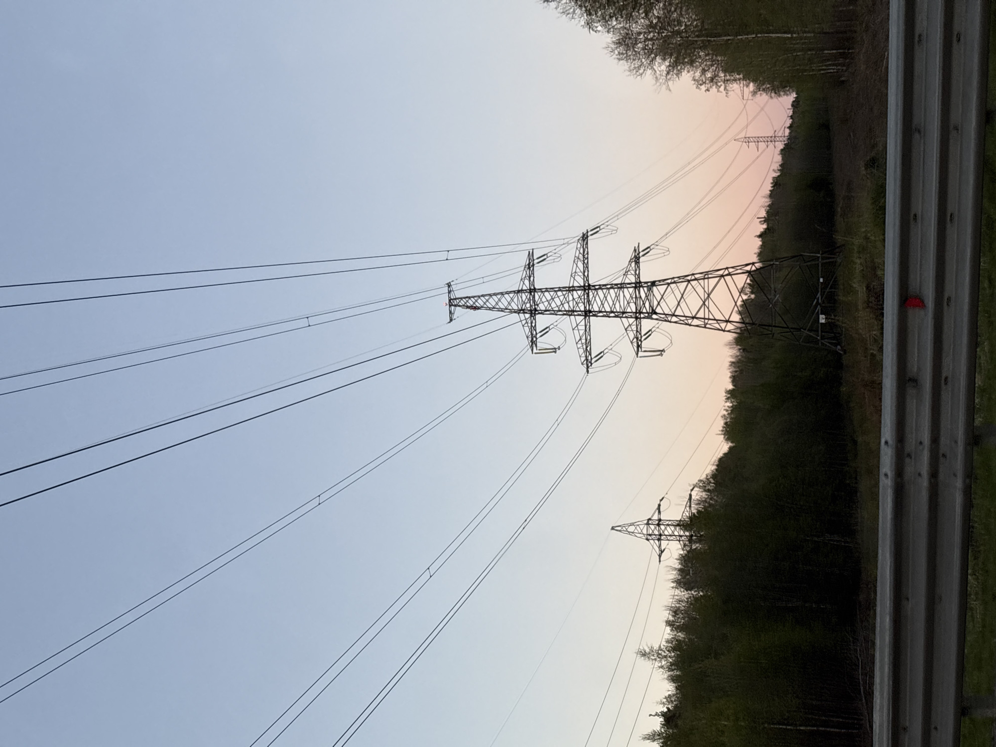 The image shows a view of electrical transmission towers and power lines in Novosaratovka, Russia. The scene is captured at what appears to be dusk, as the sky exhibits hues of light blue and pink. Trees line the side of a road which has a metal guardrail. A total of three transmission towers are visible, receding into the distance. The primary focus is a large tower in the center of the image.