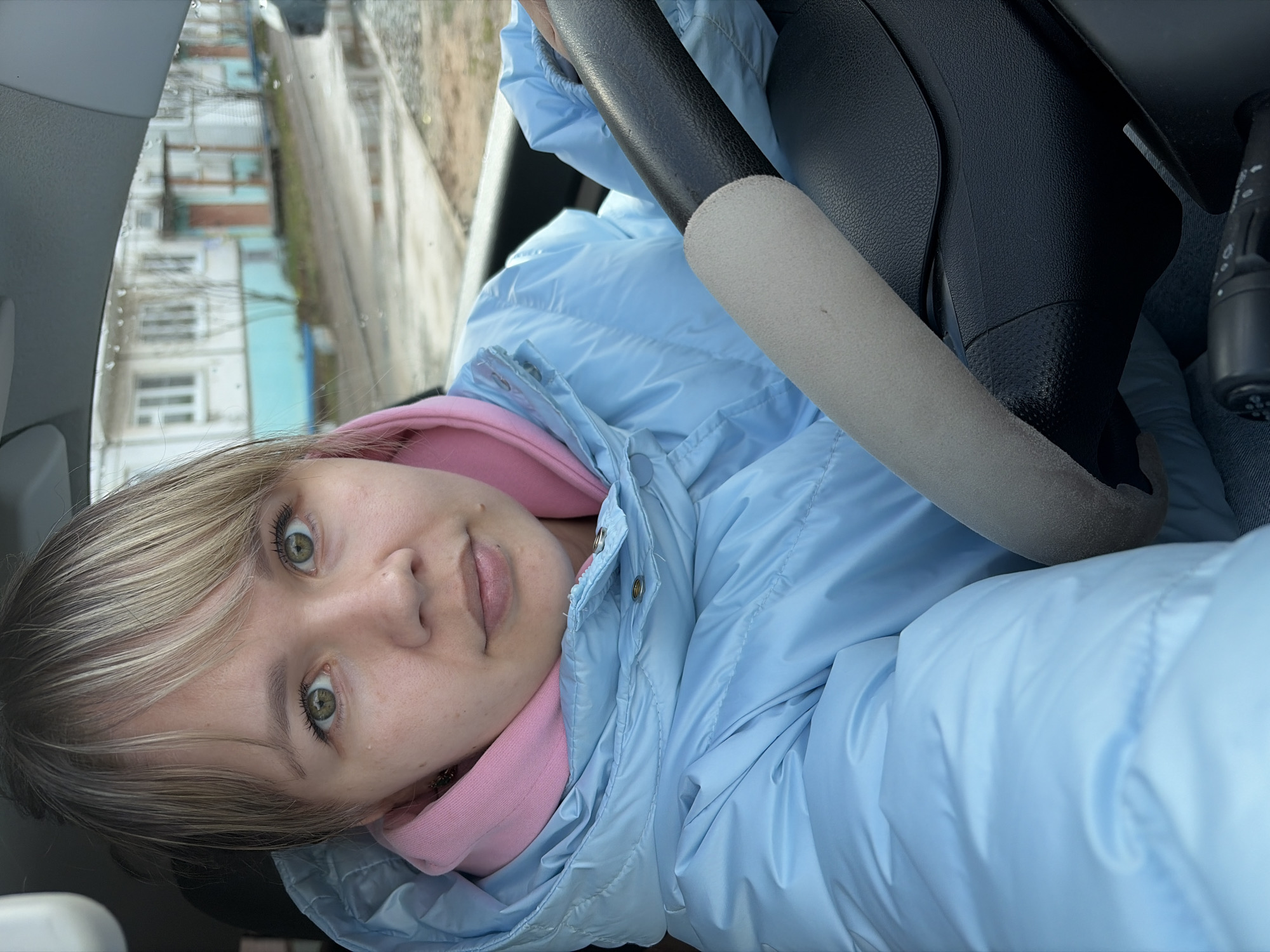 A person with light hair and green eyes is in the driver's seat of a car. The person is wearing a light blue puffy jacket over a pink hoodie. A car steering wheel is in front of them. The car is on a street. Buildings are visible outside the car window. It appears to be a cloudy day.