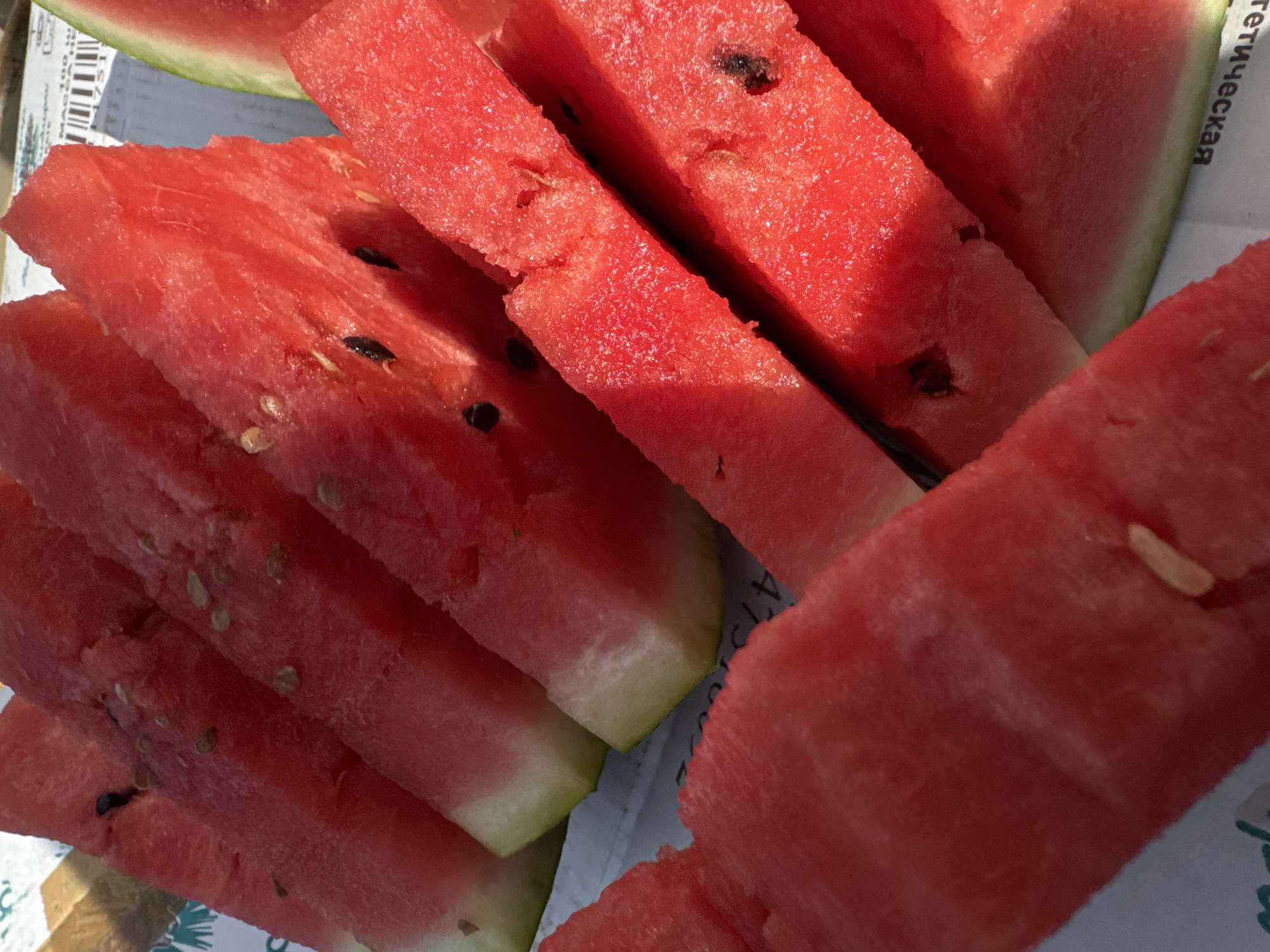 The image is a close-up, overhead shot of several slices of watermelon arranged in a fanned pattern on a white surface with some text visible. The watermelon slices are bright red with some visible seeds and a thin green rind on some pieces. The lighting suggests it is daytime, possibly outdoors, with strong shadows cast across the fruit. The text visible on the white surface is in Russian and appears to be part of some packaging or print. It is not possible to definitively determine the specific location within Chaikovsky, Russia, based on the visual information provided in this image.