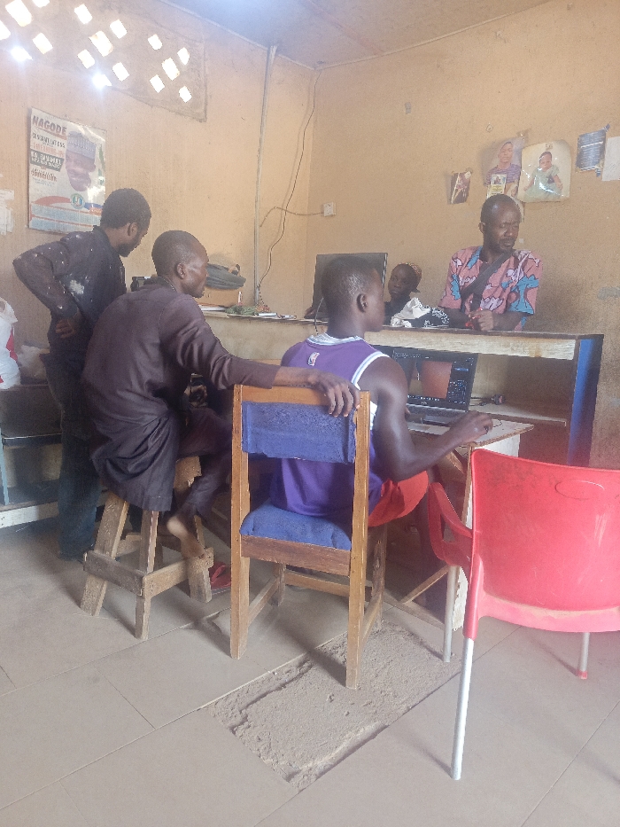 The image captures an interior scene, likely a modest commercial establishment such as a cybercafe or small office, located in Akwanga, Nigeria. Five individuals are present, all with dark complexions, engaged with a laptop computer.
In the foreground, a male subject, dressed in a purple and white NBA jersey and red shorts, is seated on a wooden chair with a blue padded seat. He is positioned with his back to the camera, actively operating a laptop on a light-colored wooden desk, with his right hand on a computer mouse.
To his immediate left, a second male subject, wearing a dark brown, long-sleeved tunic, is seated barefoot on a wooden stool, leaning forward and observing the laptop screen. His left arm rests on the back of the first subject's chair. A third male subject, dressed in a dark long-sleeved shirt and dark trousers, stands behind and to the left of the second subject, also focused on the laptop.
Behind the wooden desk, a fourth male subject with a beard, wearing a patterned short-sleeved shirt (pink, blue, white design), is seated and observing the laptop screen. Partially obscured between the first and fourth subjects, a child's head is visible, also looking towards the screen.
The room features light brown painted walls. On the left wall, a poster with "NAGODE" prominently displayed at the top, along with a male portrait, is visible. On the right wall, multiple framed or laminated photographs, including one of a woman and one of a baby, are affixed. Electrical wiring runs vertically and horizontally on the right wall. The ceiling is light-colored with square cut-outs, some of which show visible light sources. The floor is tiled with light beige square tiles, but a rectangular section in the foreground exhibits missing tiles, exposing the underlying concrete or dirt. An unoccupied red plastic chair with silver legs stands in the foreground right.