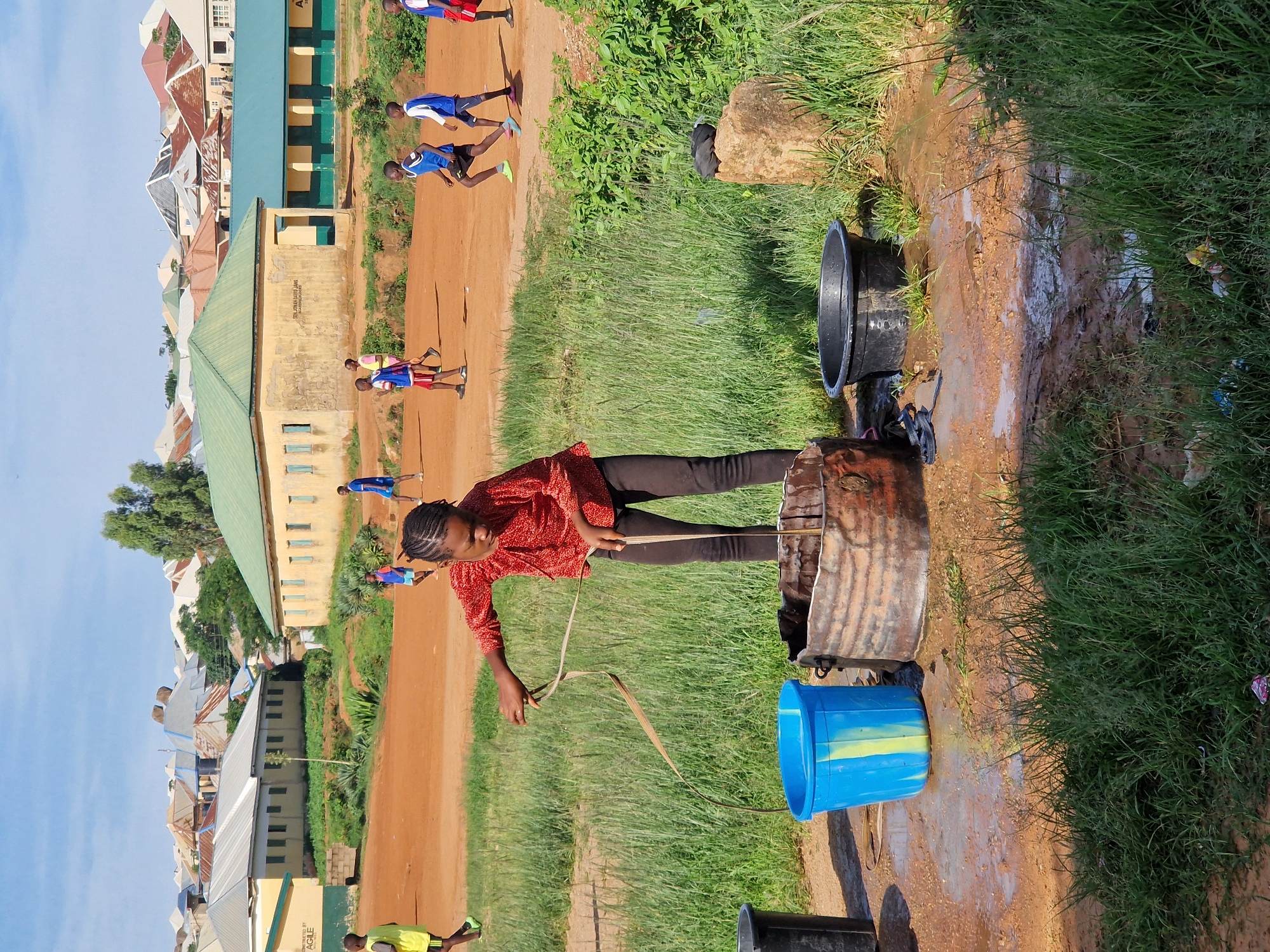 In the city of Kabong, Nigeria, under a bright blue sky, a young girl in a red, patterned shirt and black pants is drawing water from a rusty, metal drum. The drum is located on a patch of muddy ground surrounded by tall green grass. Next to the drum, a bright blue plastic bucket with a yellow stripe stands on the wet earth. The girl is holding a long, thin object, possibly a rope or a piece of cloth, with both hands as she extracts it from the drum, its end dipping into the murky water. In the background, other children, dressed in blue and red sportswear, are playing soccer on a dirt field. Beyond them, a collection of buildings with corrugated iron roofs and yellow and green painted walls are visible, characteristic of the urban landscape of Kabong. The overall scene depicts a common activity of water collection amidst a lively environment.