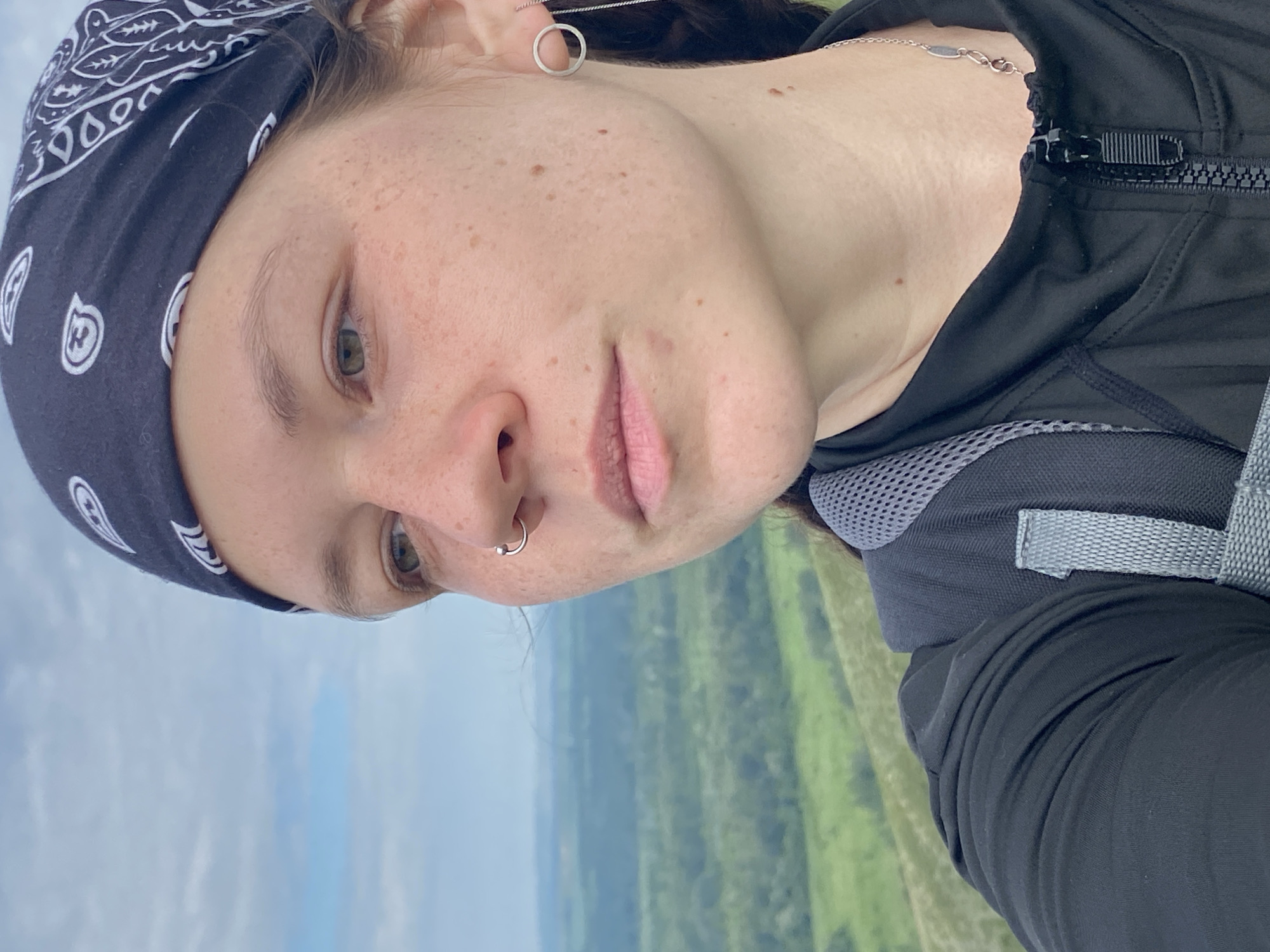 A woman wearing a black bandana with a white paisley pattern and a black zippered jacket is pictured in a close-up, over-the-shoulder shot. She has a nose ring and a silver circular earring with a chain. She is outdoors with a landscape of green hills and a cloudy sky in the background. The scene is located in Verkhnyaya Chernorechka, Russia.