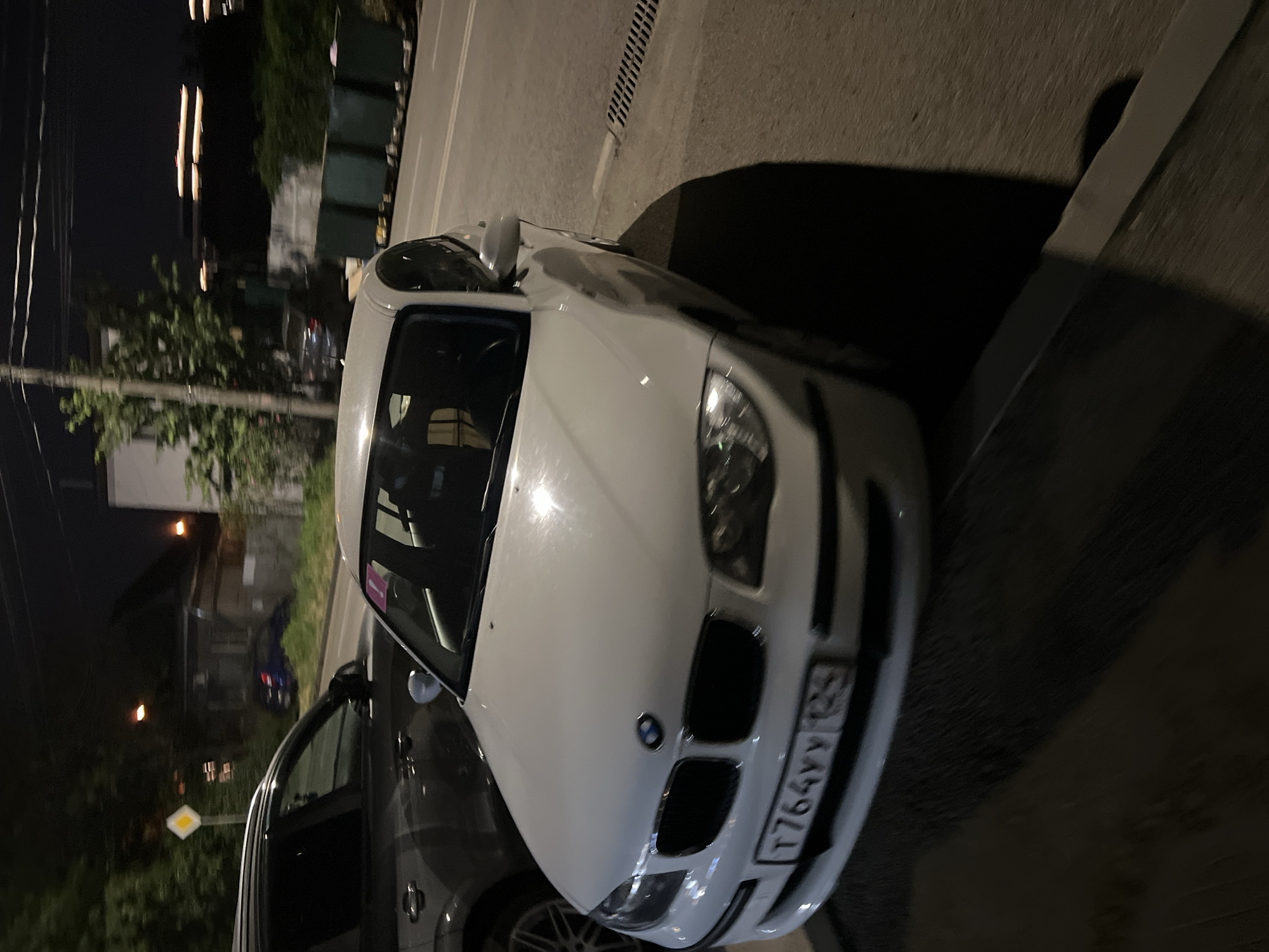 The image shows a white BMW parked on a street at night, likely in Krasnodar, Russia, based on the license plate format. The car is facing forward, and its headlights are on. The background is dark, with some trees and buildings visible, suggesting an urban or suburban setting. The street appears to be asphalt, and there's a shadow of the car cast on the ground, indicating artificial lighting or streetlights. The weather is not explicitly discernible, but the dark environment suggests it's nighttime.
The license plate of the car is visible and reads "T764YY 93". The "93" is a regional code for Krasnodar Krai, Russia. The car's front grille and emblem are clearly visible. The overall scene depicts a parked car at night, with no people or specific activities taking place. The image has a slightly tilted perspective.