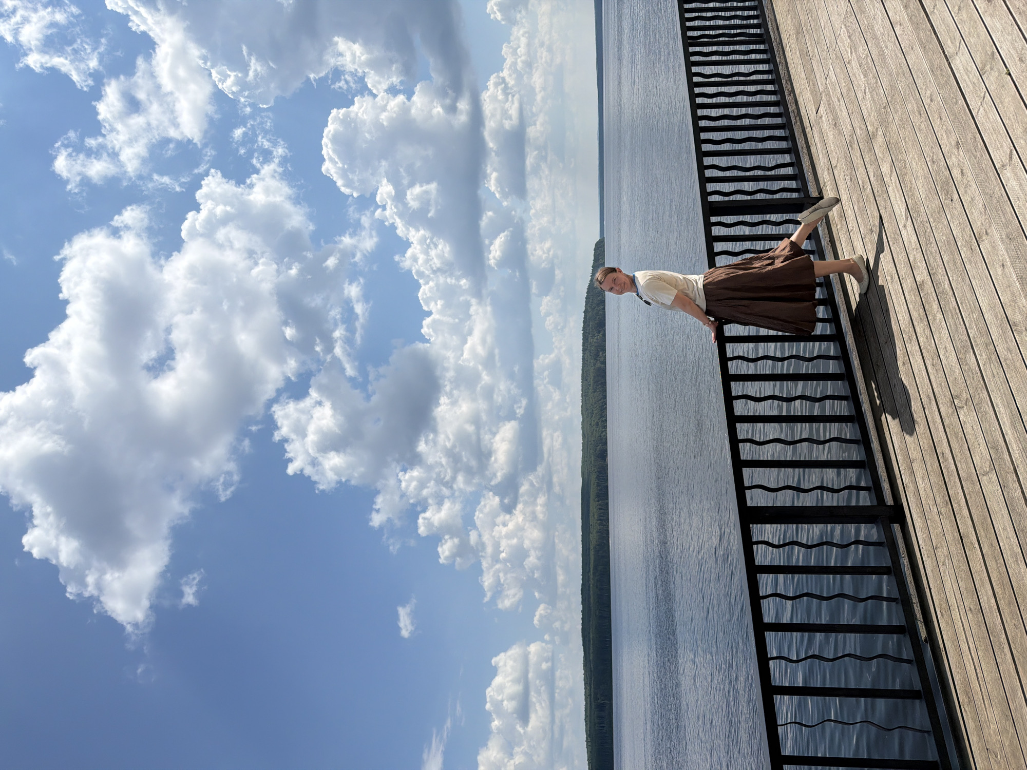 A young woman stands on a wooden pier next to a railing, with a large body of water behind her. She is looking over her shoulder at the camera with a slight smile. She is wearing a light-colored t-shirt and a long, brown, pleated skirt. The wooden pier has a weathered look and the railing is black with a wavy design. The water is calm and reflects the blue sky and white clouds. In the distance, a tree line of dark green trees is visible on the horizon. The sky is a vibrant blue with scattered, fluffy white clouds. The scene appears to be a scenic outdoor location, likely a waterfront park or recreational area. This location is known to be the embankment in the city of Chaikovsky, Russia.