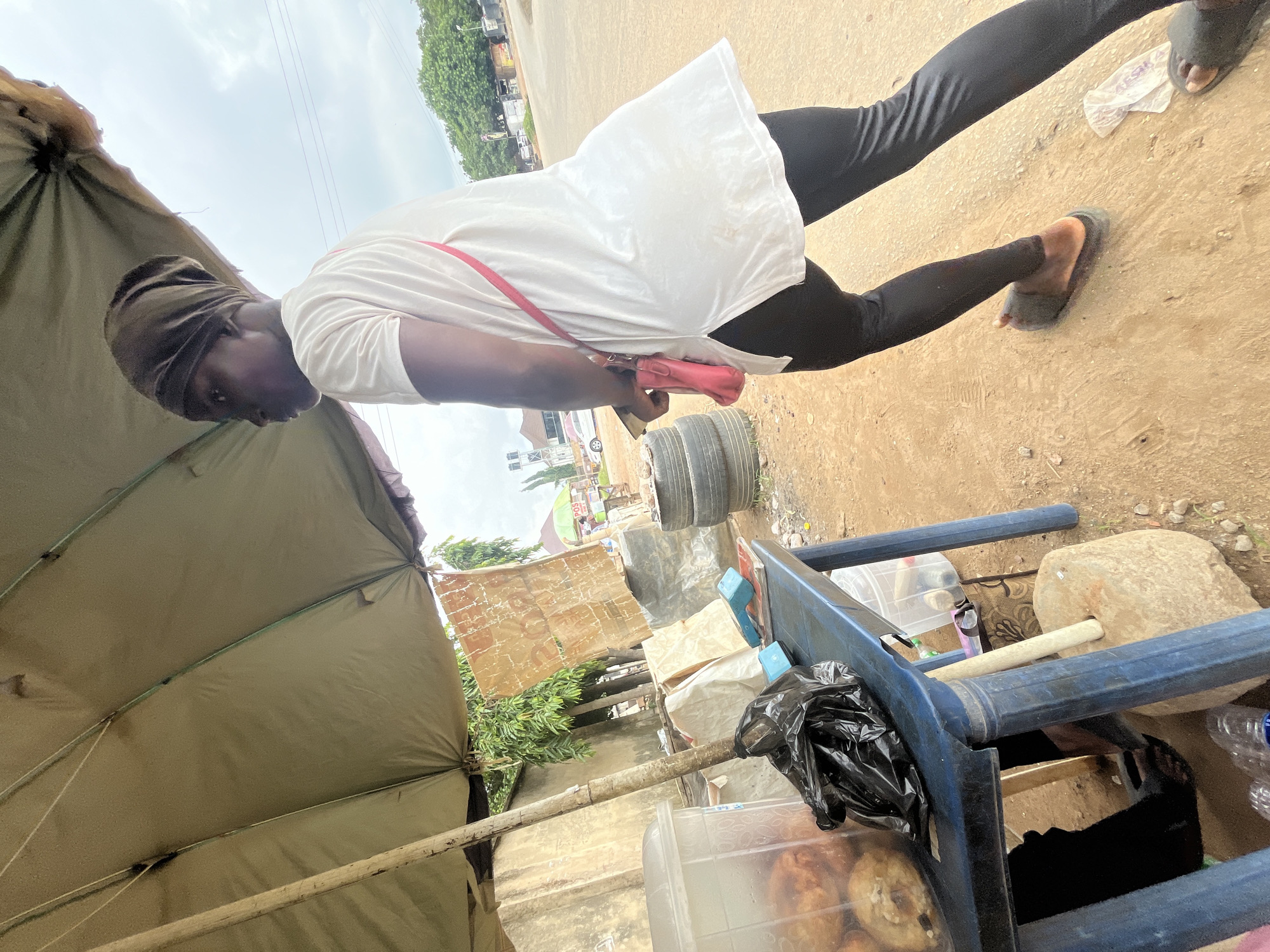 A person, likely female due to the attire, stands on a dirt path under a large, olive-green awning, possibly a makeshift stall. The person wears a white t-shirt and black leggings, with a brown headscarf and a small pink bag slung across their chest. Their body is angled away from the camera, looking towards the right. In the foreground, a blue plastic table is laden with items, including a clear container filled with what appears to be fried food, a black plastic bag, and other miscellaneous objects. Behind the table, stacked tires and a concrete wall are visible. The background shows a street with sparse vegetation, distant buildings, and utility poles, suggesting an outdoor urban or peri-urban setting in Abuja, Nigeria. The lighting suggests an overcast day.