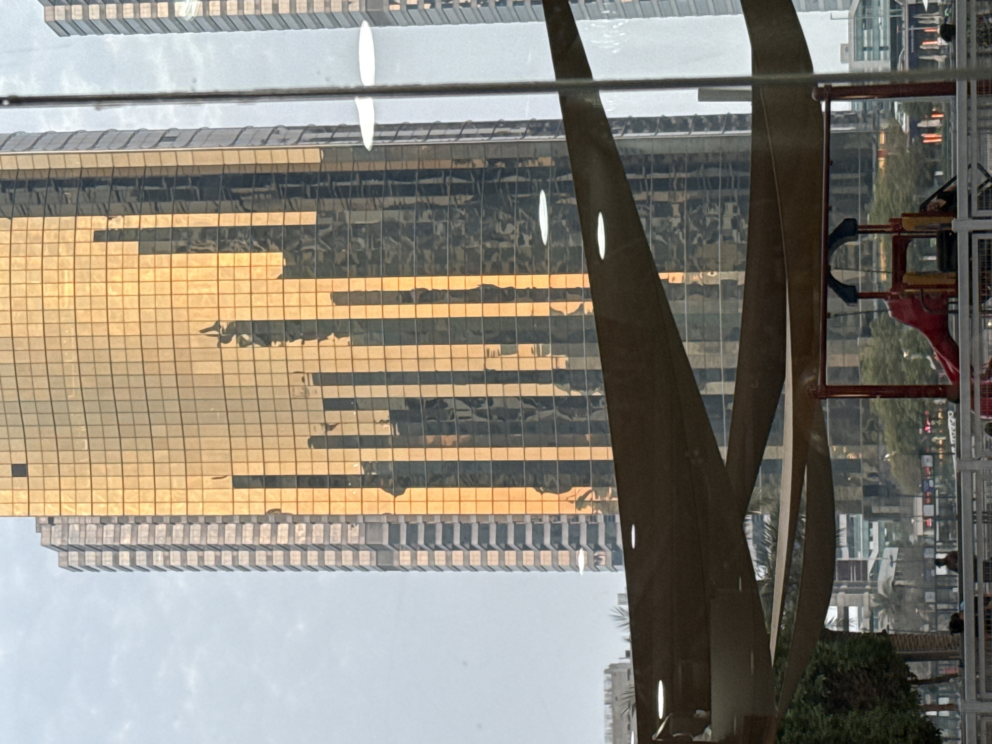 The image captures a view from a shaded outdoor area, possibly a park or a public space, looking towards a dense urban cityscape. Dominating the background are tall, modern skyscrapers, characteristic of Dubai. The most prominent building is a massive skyscraper clad in gold-colored glass, reflecting the sky and surrounding structures in a distorted, abstract pattern. To its left, another skyscraper, clad in light-colored, segmented panels, stands tall. In the foreground, a large, brown fabric awning stretches across the scene, supported by structures not fully visible. Beneath the awning, a children's playground with a red slide and yellow structure is partially visible. The environment suggests a daytime setting, possibly late afternoon, given the warm glow of the sun on the gold-tinted building. The overall impression is one of a vibrant, contemporary urban environment, juxtaposing natural elements like shade canopies with the imposing architecture of Dubai.