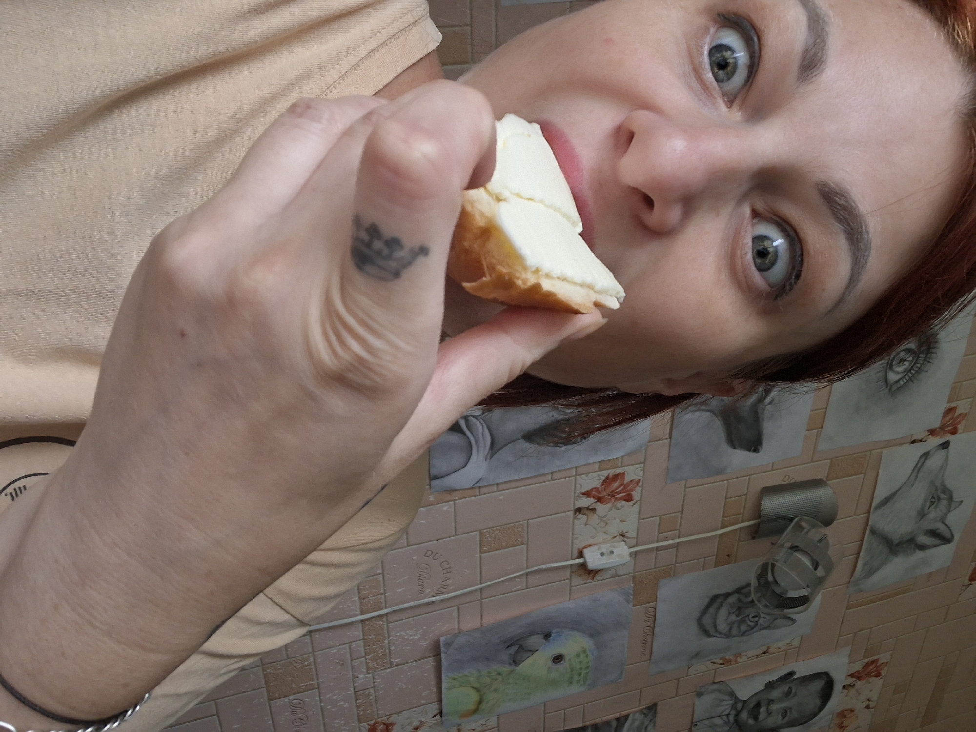 A woman with reddish-brown hair is shown from the chest up, her eyes wide with surprise, and she is about to eat a piece of white food. She is wearing a light peach-colored t-shirt. Behind her is a wall tiled with light pink and beige squares. Several framed drawings are hanging on the wall, including a portrait of a fox, a cat, a child, an eye, a dog's head, and a parrot. A clear plastic light fixture is attached to the wall. The environment is indoors, likely a home, and given the artworks, it could be an artist's studio or a living space decorated with art. There is no identifiable information to place this image in Myadzyel, Belarus.