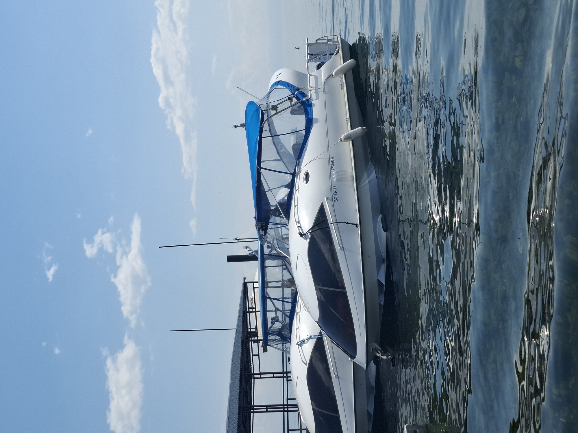 Two white boats with blue canopies are docked on a calm body of water. The boats are modern in design, with sleek, aerodynamic hulls and large, tinted windows. The boat on the right has "BC-23-102 АРКАНУМ" and "looker 31" written on its side. The boats are tied to a dock with a roof structure, suggesting they are in a marina or at a ferry terminal. The water is clear and reflects the sky and the boats. The sky is blue with scattered white clouds, and the sun appears to be shining. The image captures a serene moment in a waterfront location, likely Listvyanka, Russia, a popular tourist destination on the shores of Lake Baikal.