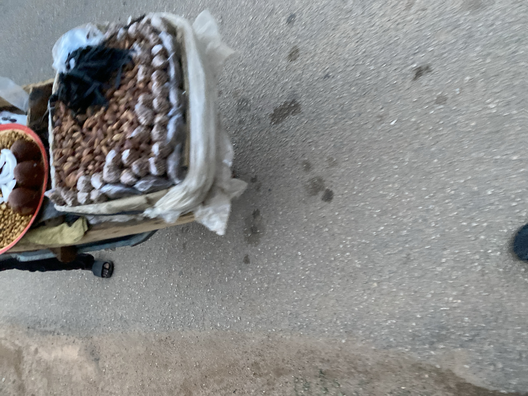 The image displays a ground-level view of a street vendor's stall, likely situated on a paved road or market area in Jos, Nigeria. A wooden stand or cart supports various snack items. The main offering is a large quantity of light brown, irregularly shaped items, strongly resembling roasted groundnuts (peanuts), filling a container. Some darker items, possibly other nuts or dried fruit, are mixed in, and a black, undefined material (perhaps a cloth) rests on top of some nuts. White plastic sheeting or bags are draped around the display.
To the left of the main container, a smaller red basket holds several dark brown, uniformly round items, which could be local sweets or another type of snack. A small piece of white material is also visible within this basket. The stall is positioned on a grey, textured asphalt or concrete surface, showing some darker stains. A human presence is indicated by a foot, clad in a black open-toed sandal, visible beneath the stand on the lower left, suggesting the vendor is standing by. The scene is illuminated by daylight. No visible text is present.