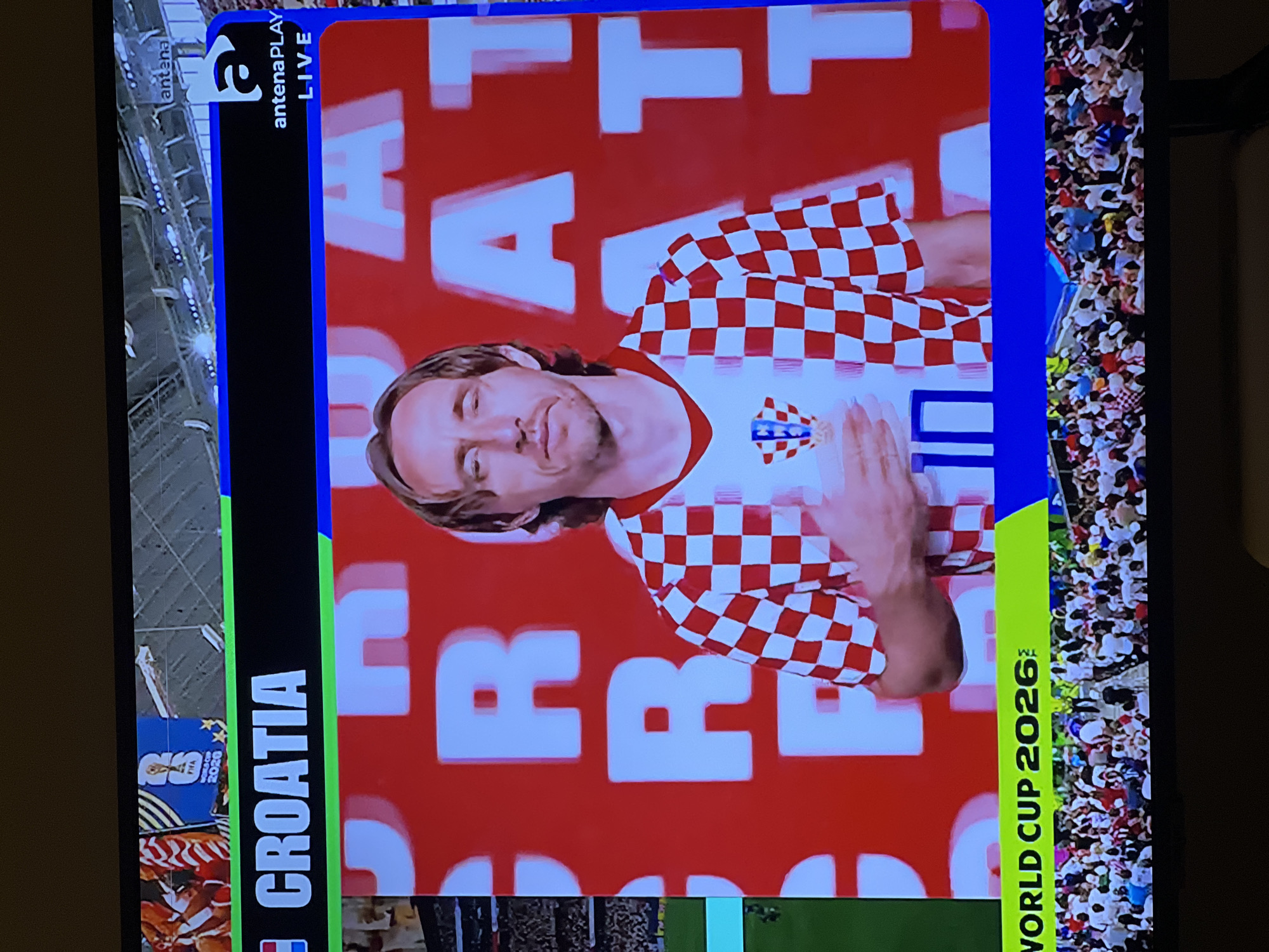 The image shows a television screen displaying a broadcast related to Croatia and the World Cup. A man, likely a footballer, is shown prominently in the center wearing a red and white checkered jersey, the national colors of Croatia. He has his hand on his chest. Behind him, large white letters spell out "CROATIA" and "WORLD CUP 2026". The broadcast also shows logos for "antena PLAY LIVE" and "FIFA WORLD CUP 2020". The bottom of the screen shows a crowd of people watching the broadcast. There is no indication in the image that this scene is located in Florești, Romania.