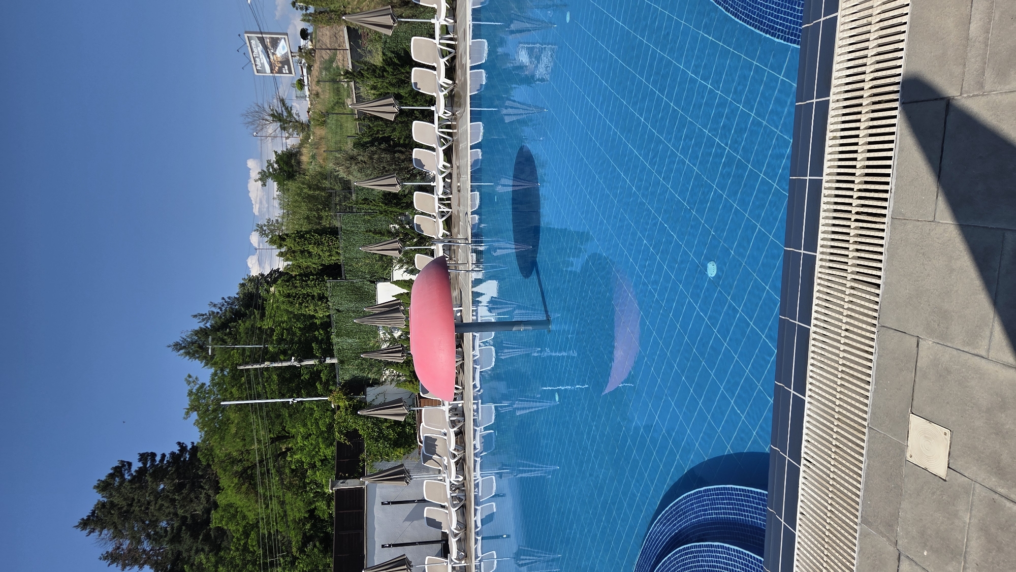 An outdoor swimming pool scene is depicted on a clear day with a bright blue sky. The pool is filled with bright blue water, featuring a grid pattern of tiles on its bottom. In the center of the pool, a red umbrella-like structure stands on a pole, casting a reflection on the water's surface. Around the pool's edge, lounge chairs and closed umbrellas are arranged on a tiled patio. The background consists of lush green trees and vegetation, with a fence and a building visible behind them. A billboard with text and an image is situated on a hill in the distance. The foreground shows the edge of the pool and the surrounding tiled walkway.
