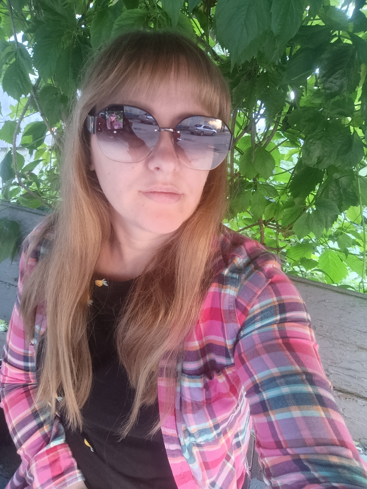 The image is a close-up selfie of a woman with blonde hair and bangs. She is wearing large, round sunglasses that reflect a street scene with cars. She has on a black t-shirt with a yellow floral pattern and a pink, purple, and blue plaid flannel shirt over it. The woman is sitting outdoors, with green foliage and branches visible behind her. A wooden bench is also partially visible. The lighting suggests it is daytime, possibly late afternoon, with dappled sunlight filtering through the leaves. The overall mood appears casual and relaxed. There is no visible text in the image.