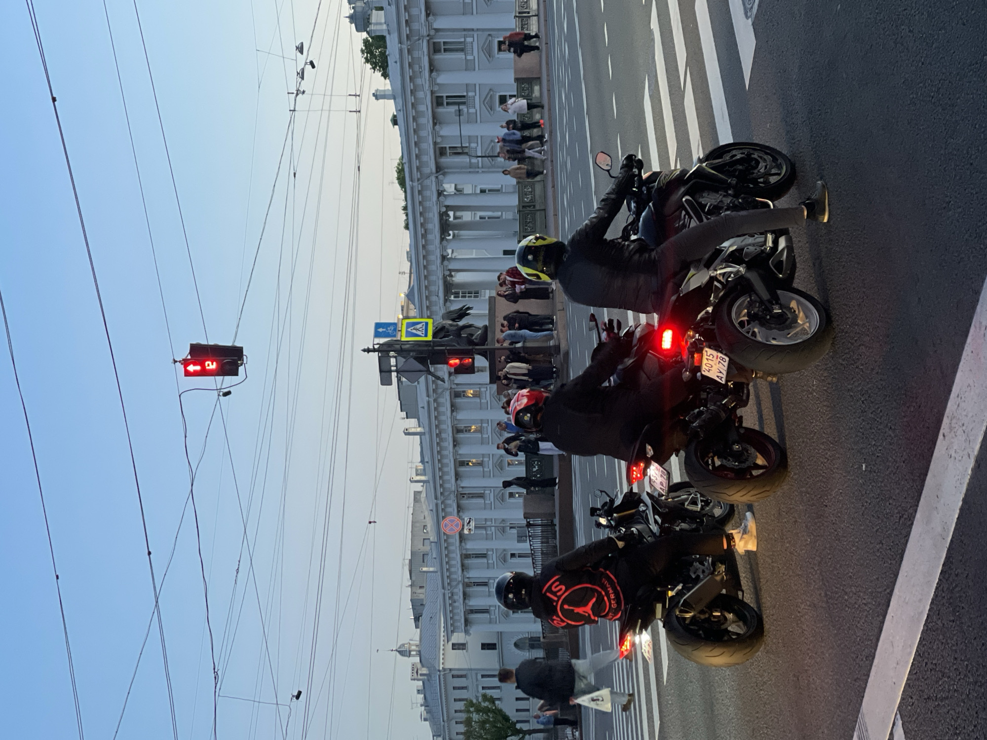 The image captures an urban street scene in Saint Petersburg, Russia, during what appears to be late afternoon or early evening. The sky is light blue and clear, suggesting fair weather.
In the foreground, three motorcyclists are stopped at a red traffic light, positioned on a marked crosswalk. The traffic light, hanging from a web of overhead wires, clearly displays a red signal with a left-turn arrow illuminated. The motorcyclists are wearing helmets and protective gear. The rider closest to the bottom of the frame has a black jacket with a prominent red and black logo on the back, partially visible as "S GERMAIN" and a Jumpman (Jordan) symbol, likely indicating a Paris Saint-Germain collaboration. Another motorcycle rider, in the middle, wears a black jacket and a yellow helmet. The third rider is further back.
On the street, white zebra crossing stripes are visible, along with other road markings. The license plate of the middle motorcycle is partially visible, displaying "4015 APJ 78," where "78" is the region code for Saint Petersburg, confirming the location.
To the left of the road, a crowd of pedestrians is gathered on a sidewalk next to grand, classical-style buildings featuring white columns and ornate details, characteristic of Saint Petersburg architecture. Visible signs include a blue square sign with a bicycle and a left arrow, and a yellow square sign with a black left arrow. Another circular red sign with a diagonal line (no stopping/standing) is also present near the buildings. Numerous overhead wires crisscross the sky, suggesting the presence of trams or trolleybuses.