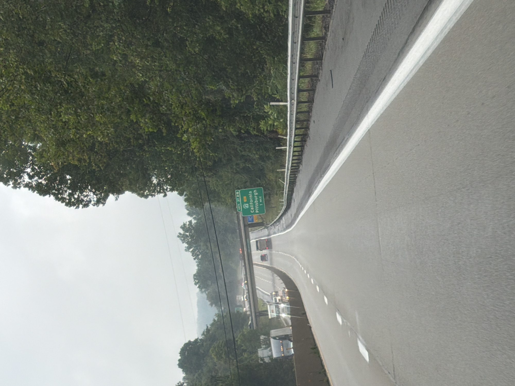 This image depicts a highway scene in Fallowfield Township, United States of America, under an overcast sky. The perspective is from a vehicle traveling on the road, looking forward. A green highway sign indicates "Exits 37 A-B California Pittsburgh 1 MILE". The road is asphalt, with white lane markings. On the right side of the road, a metal guardrail is visible. Trees and foliage line the sides of the highway. In the distance, an overpass is seen with traffic on it. Several vehicles, including trucks and cars, are on the highway. The overall environment appears to be a rural or semi-rural highway setting, likely during the daytime given the visibility.