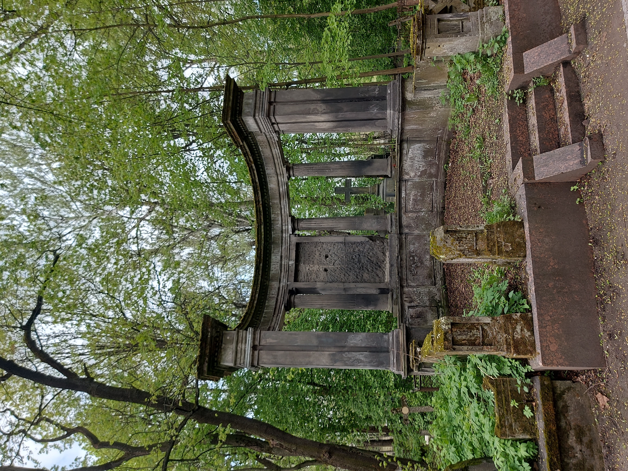 The image depicts a deteriorated stone structure in a cemetery in Saint Petersburg, Russia. The structure is dark gray, possibly made of granite, and features an arch supported by pillars. The architecture suggests a memorial or entrance to a burial plot. The structure is heavily weathered, with moss and plant growth covering parts of the stone. Behind the structure, one can see other gravestones and crosses, further indicating a cemetery setting. The surrounding environment includes trees with green foliage, suggesting a park-like setting for the cemetery. To the right of the main structure, a set of steps leads up to a higher level, also made of stone. The overall scene has a somber and historical ambiance.